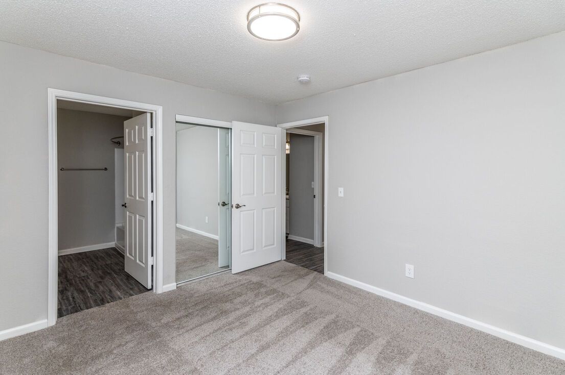 Bedroom with three white doors: closet, bathroom, and another room. Gray walls, carpet, and ceiling light.