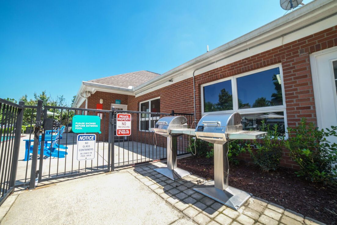 Two stainless steel grills next to a brick building, gate to a pool area with blue chairs.