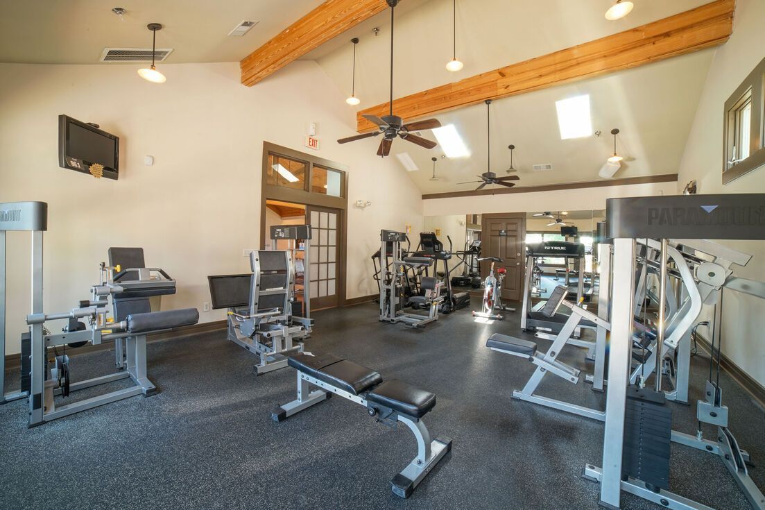 Gym interior with various exercise machines, wood beams, ceiling fans, and natural light.