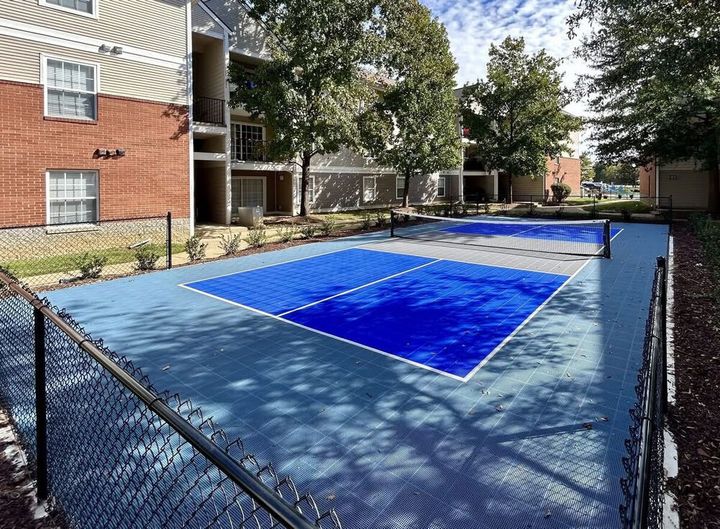 Pickleball courts with blue and light blue surface, surrounded by fence and buildings.
