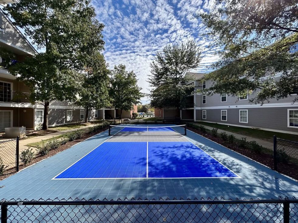 Pickleball court with blue and gray surfaces, bordered by buildings and trees under a cloudy sky.