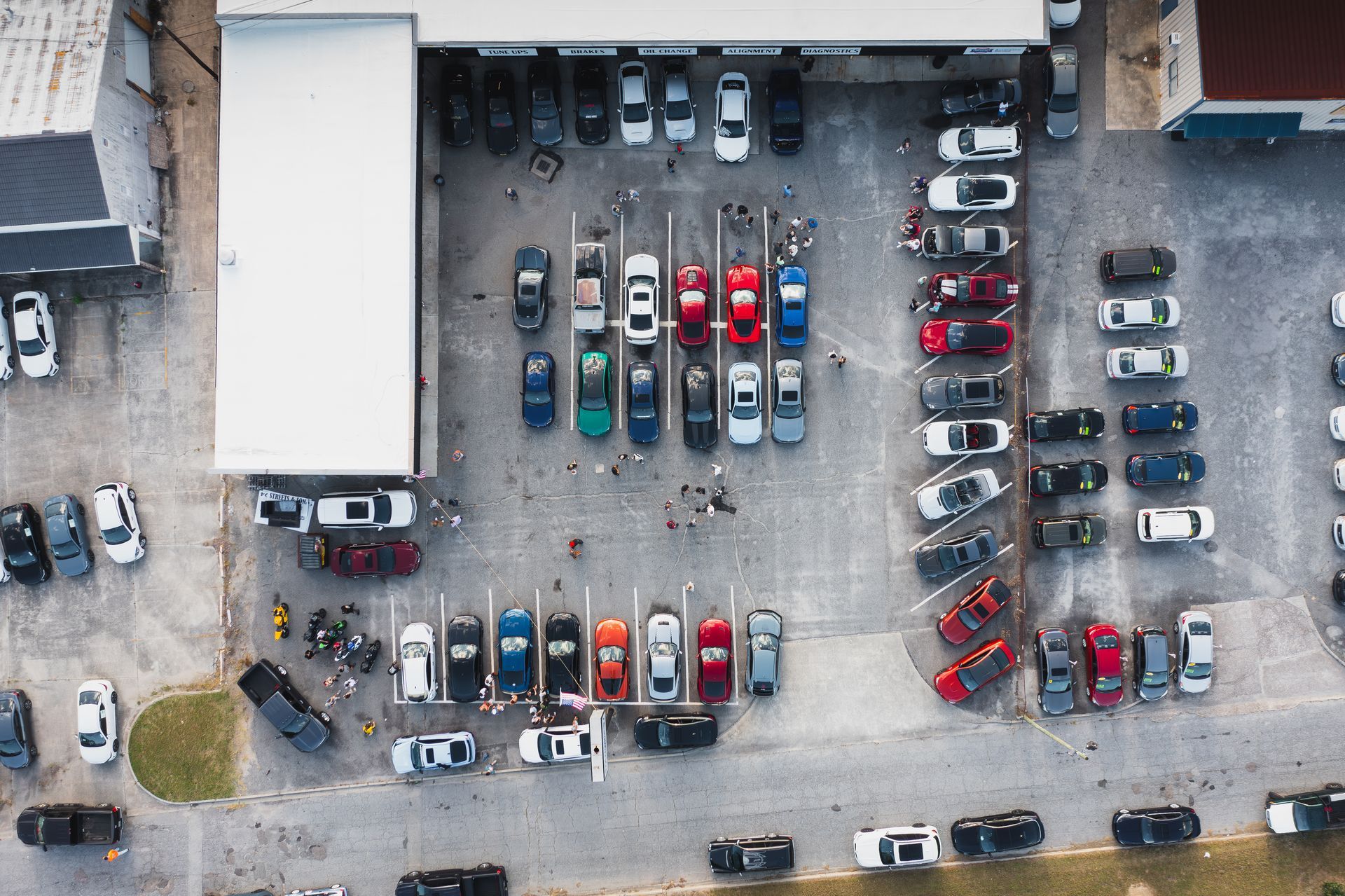 Aerial view of a car show with rows of various colored cars parked in a lot. People gather.