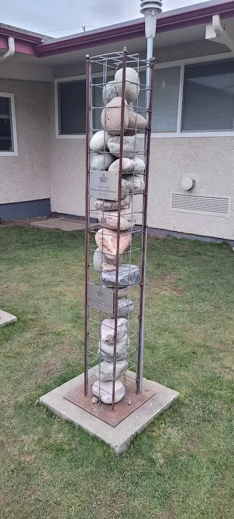 A tall metal sculpture filled with various sized stones sits on a concrete base in a grassy yard.