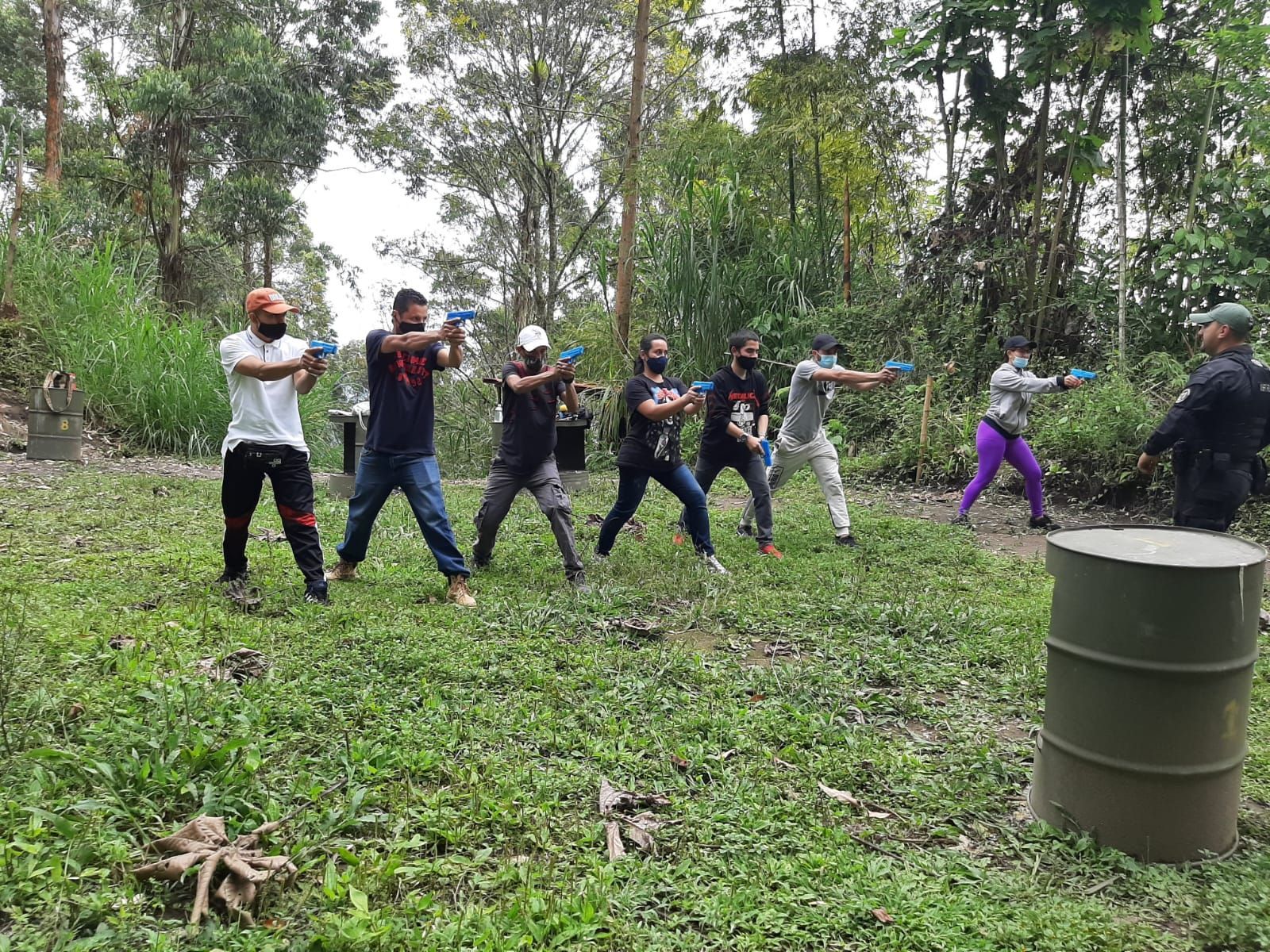 La gente practica apuntar con pistolas de juguete en un entorno al aire libre; se ven árboles, pasto alto y un barril.