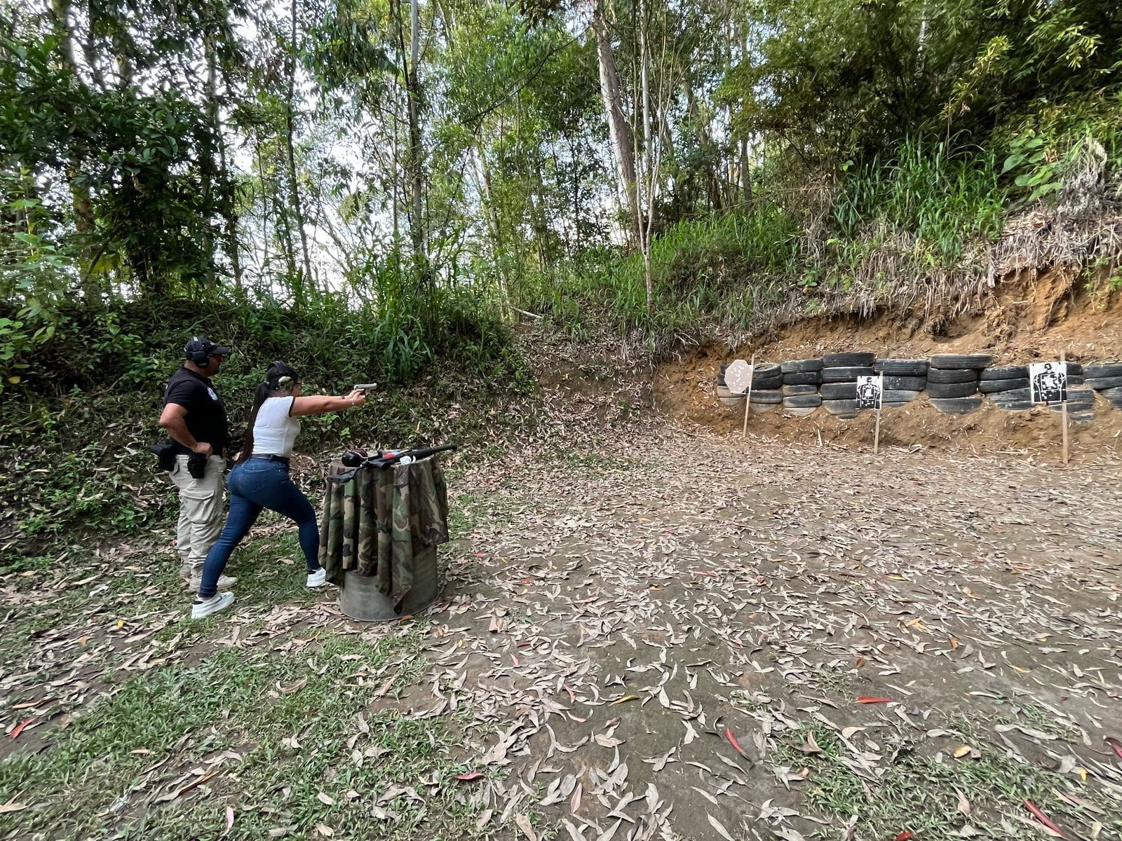 Mujer apuntando con una pistola a un objetivo en un campo de tiro, con un instructor a su lado, rodeada de árboles.