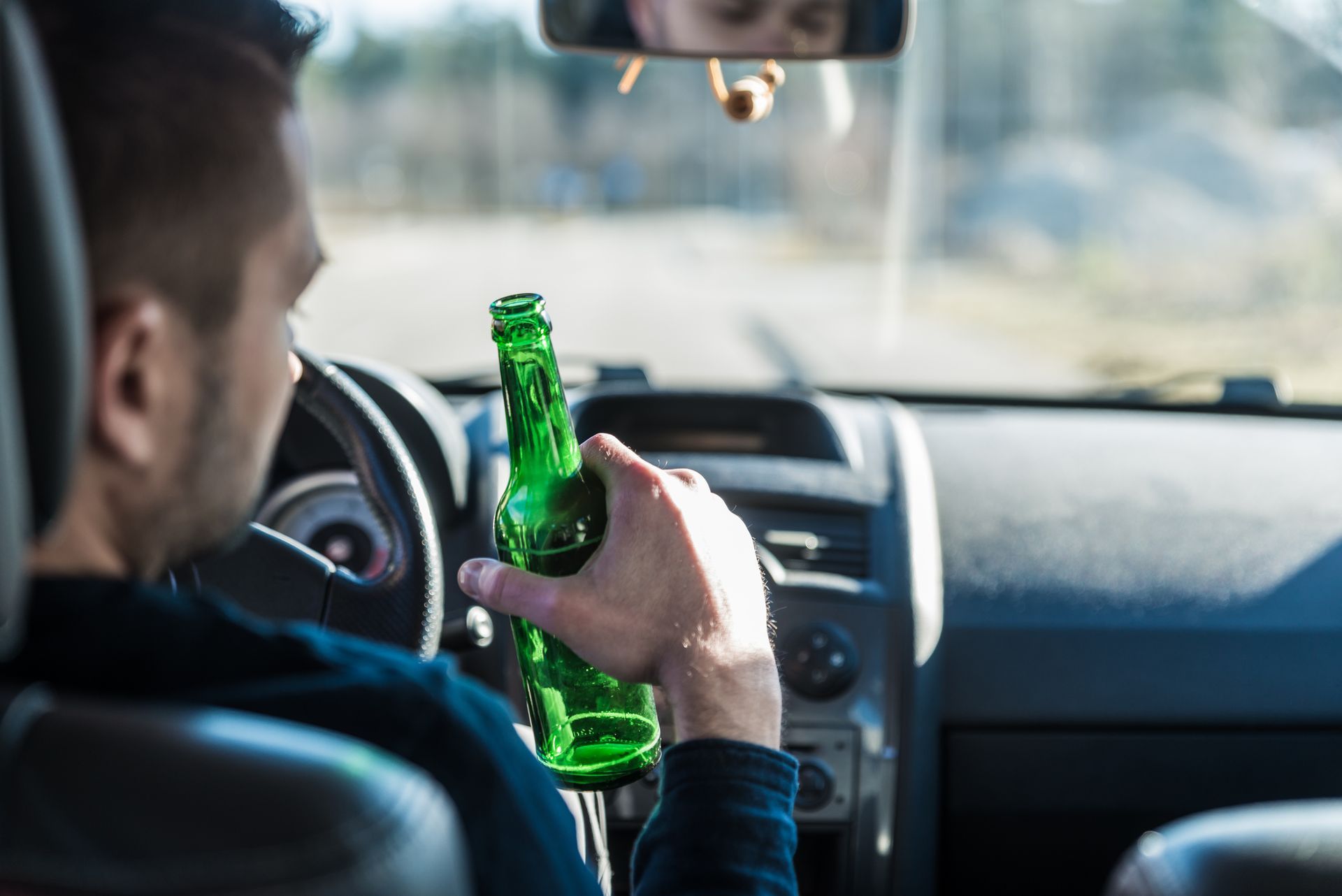 A driver holding a green beer bottle inside a car, with the steering wheel visible.