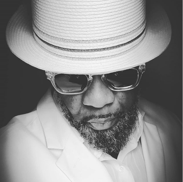 Man in white hat, sunglasses, and suit, looking down.