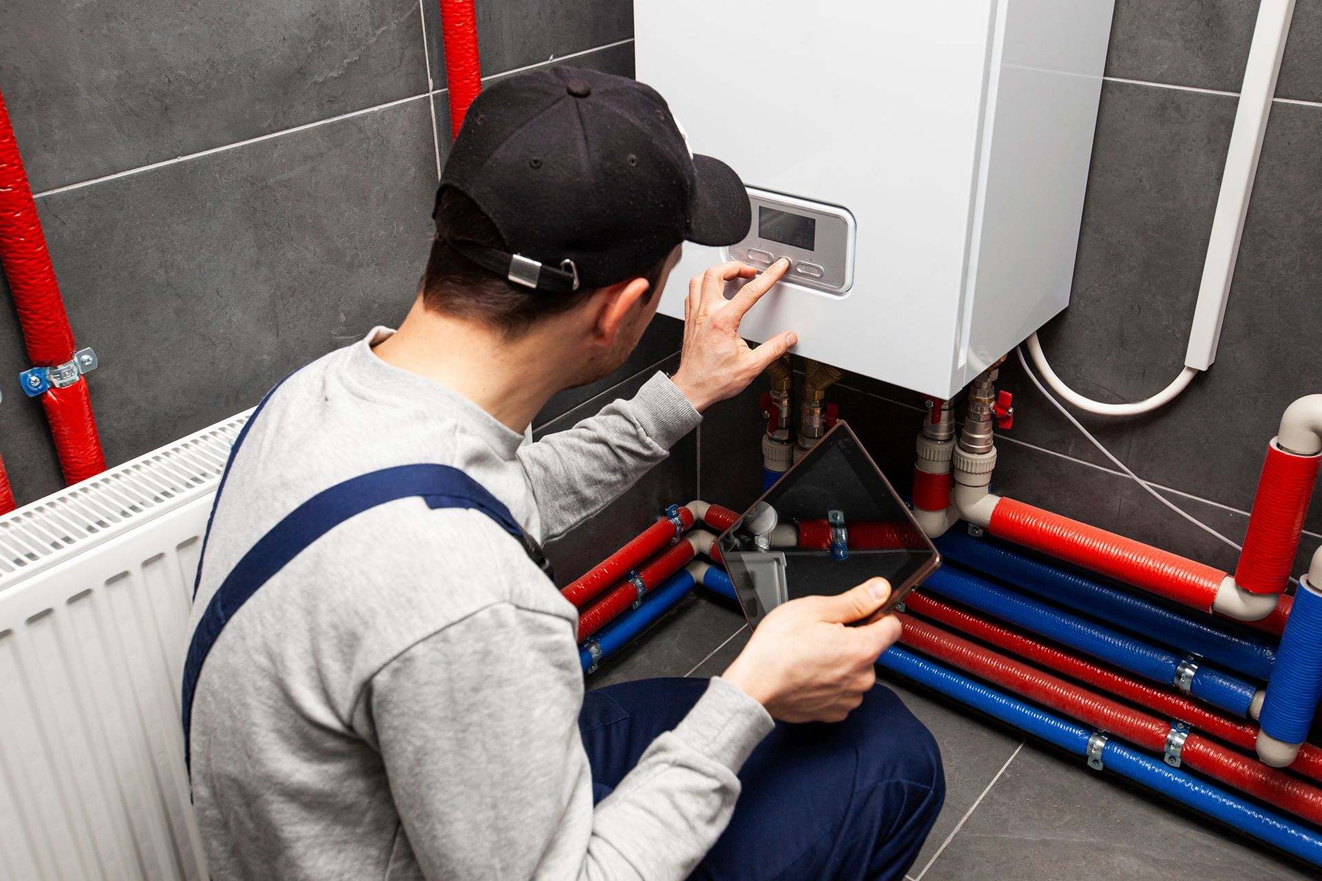 A man is kneeling down in front of a boiler and looking at a tablet.