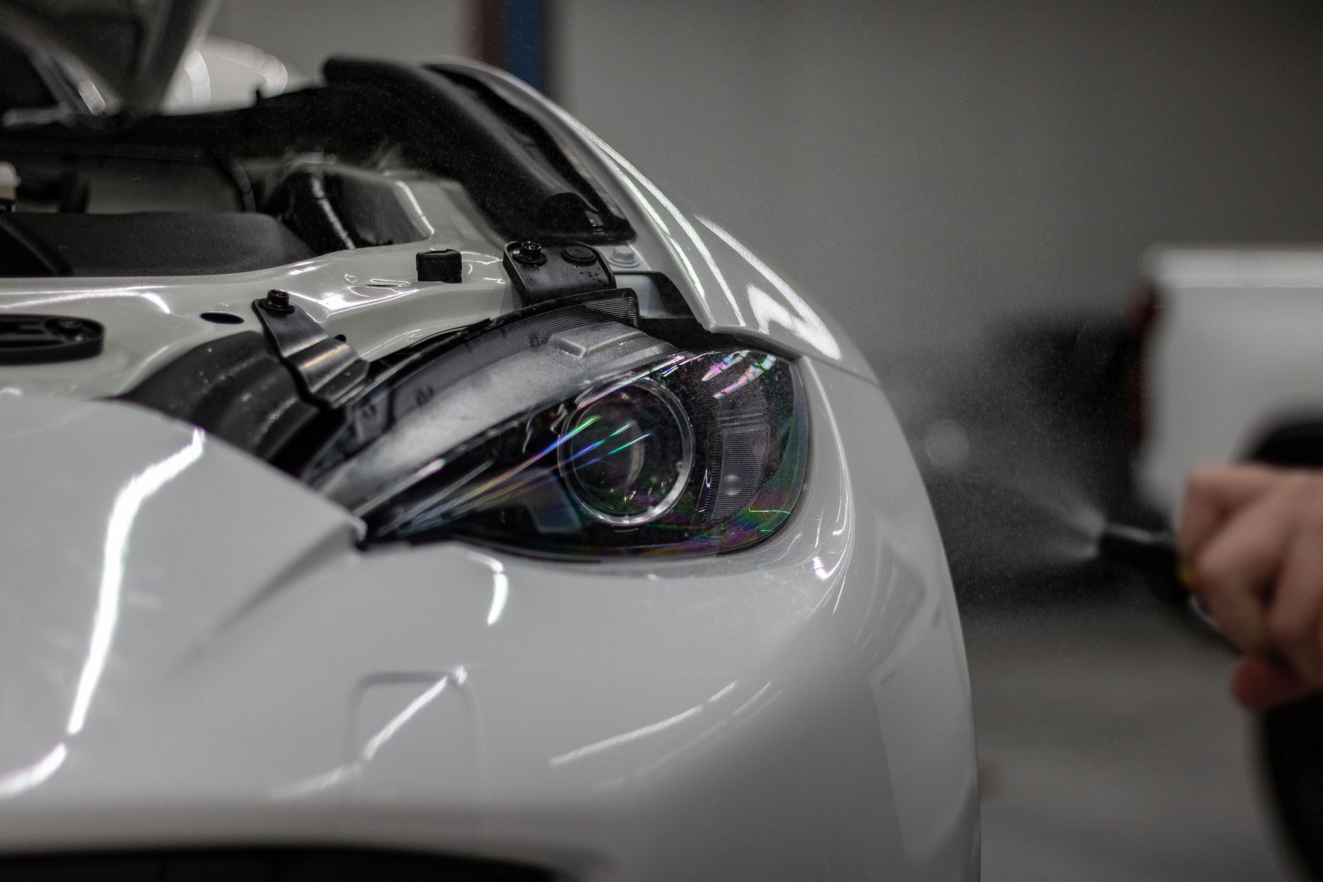 A person sprays a mist onto the white, open hood and headlight of a car in a workshop.