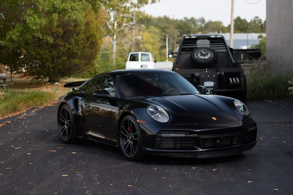 A sleek, black Porsche 911 parked on an asphalt driveway next to a black utility vehicle.