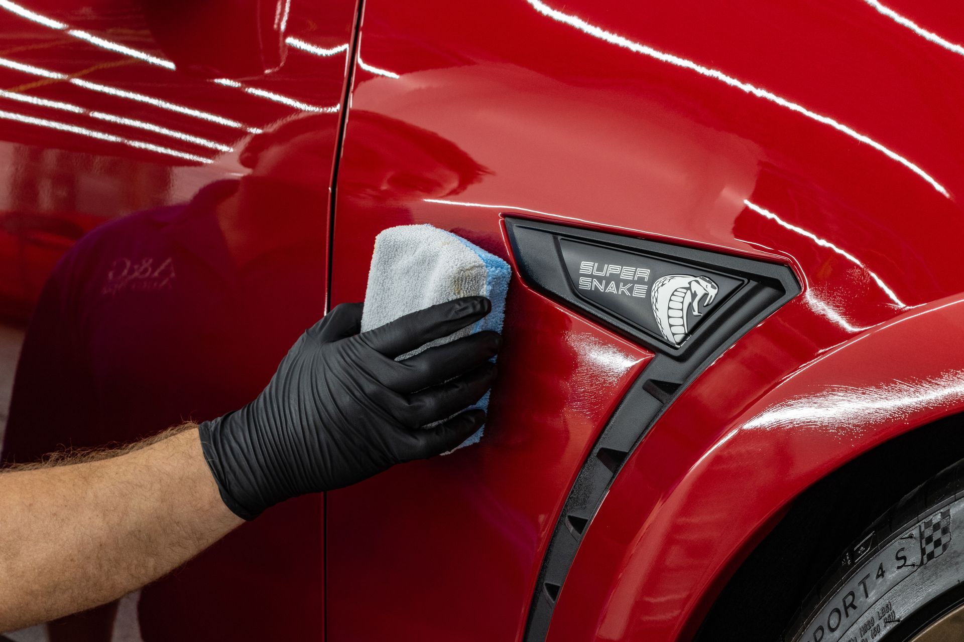 A gloved hand uses a foam pad to apply a ceramic coating to the red fender of a Shelby Super Snake car.