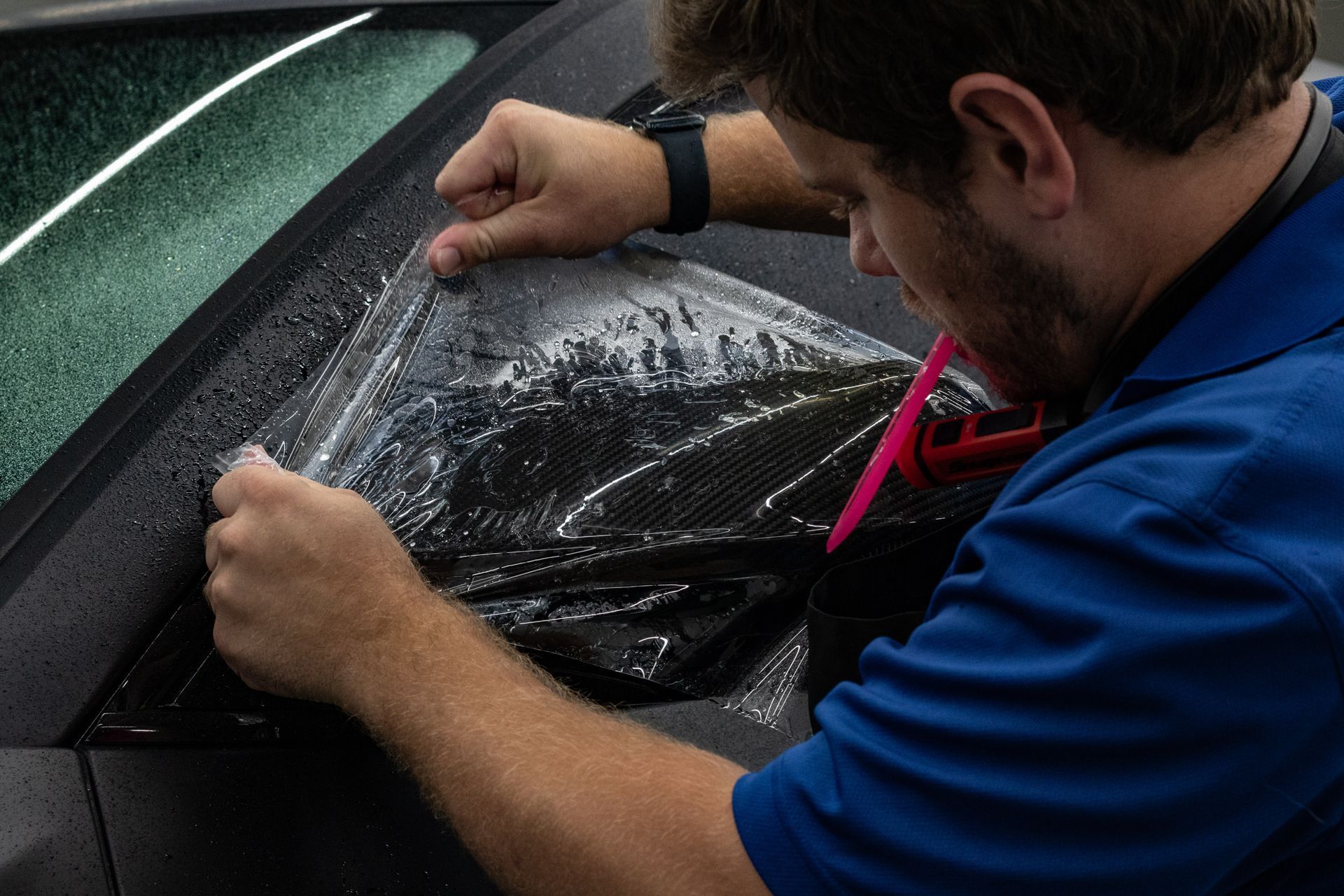A man in a blue shirt is applying a clear film to a car window.