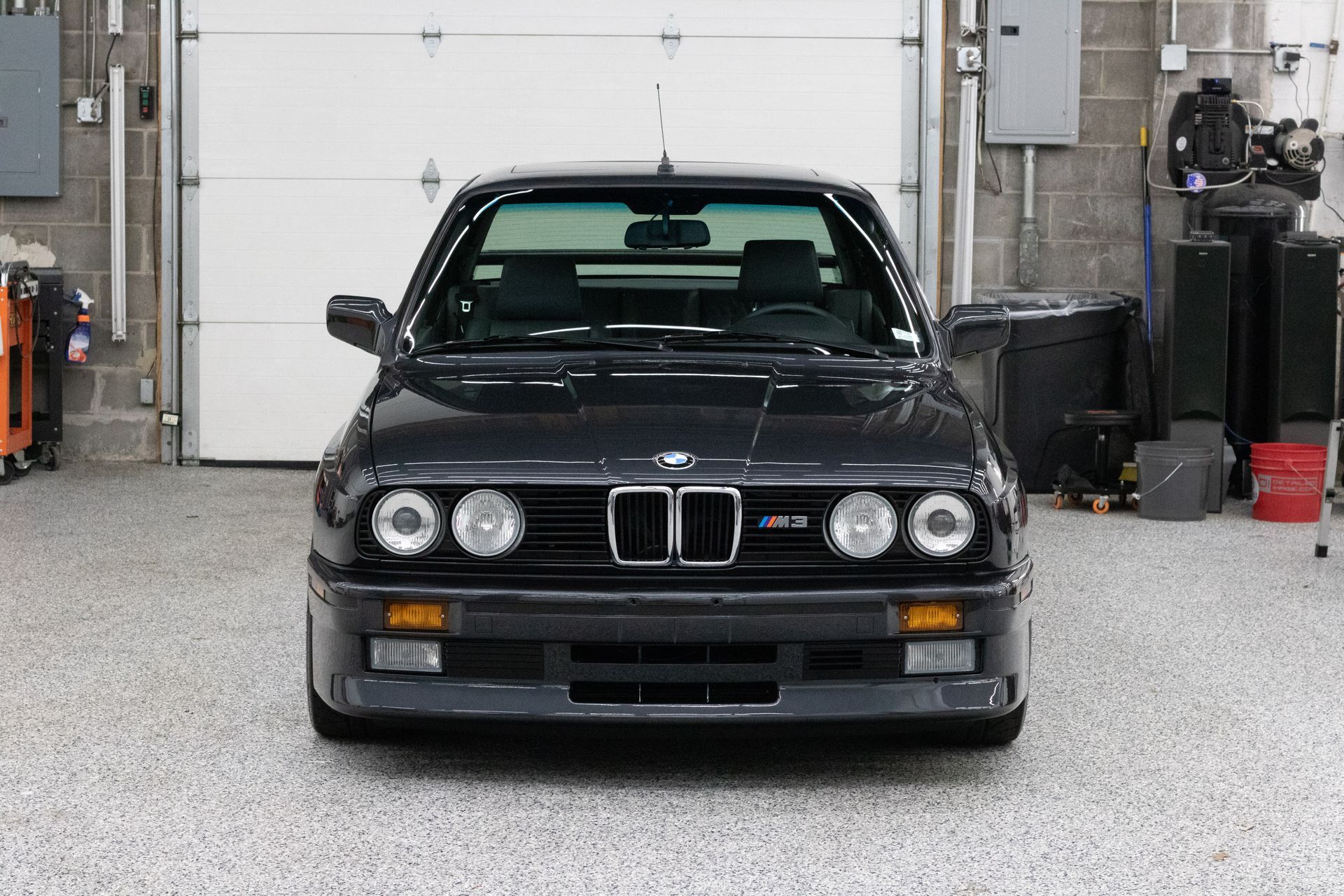 Dark gray BMW E30 M3 parked in a garage, front view, showing the car's iconic headlights.