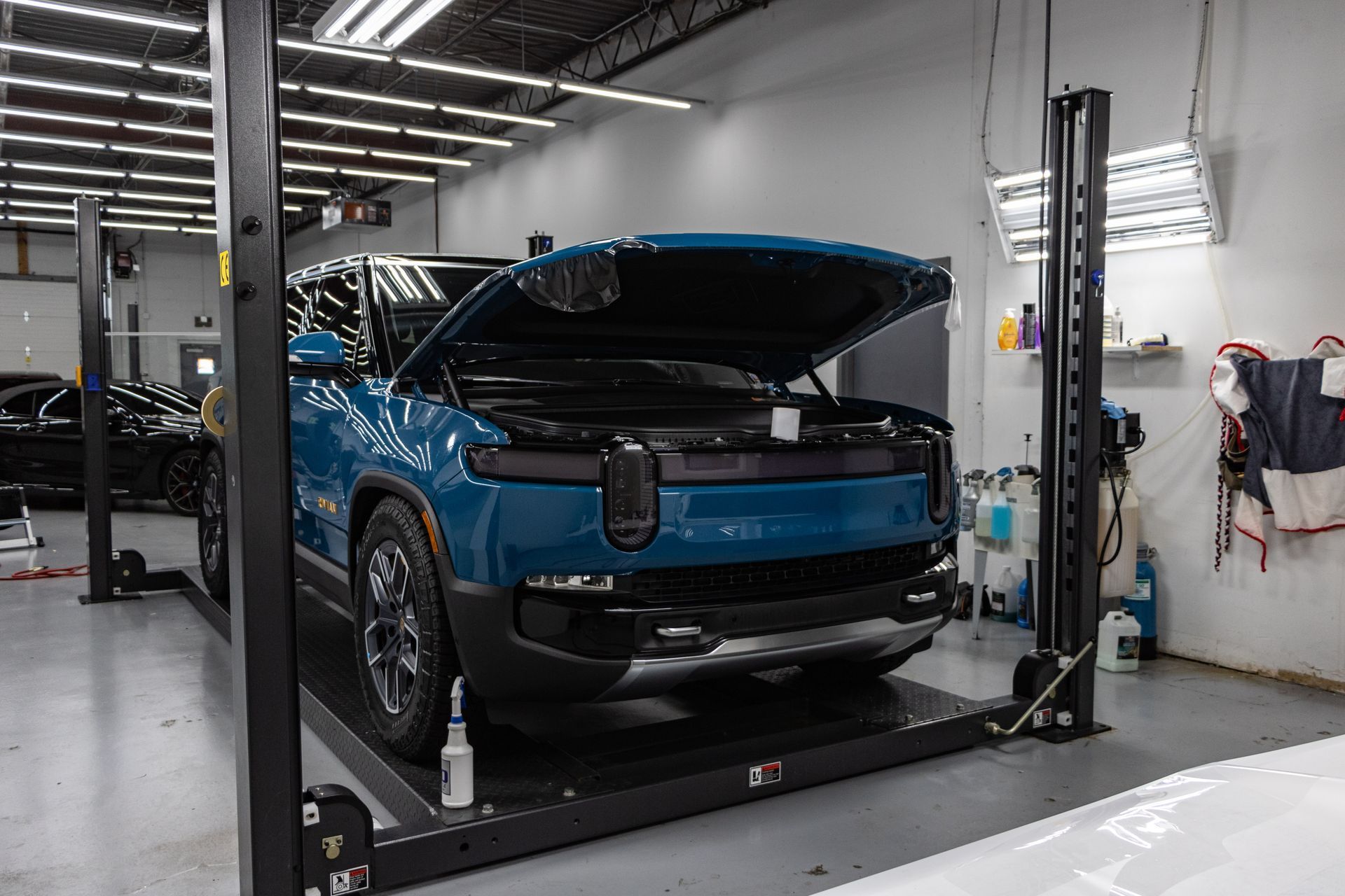 A blue car with its hood up is on a lift in a garage.