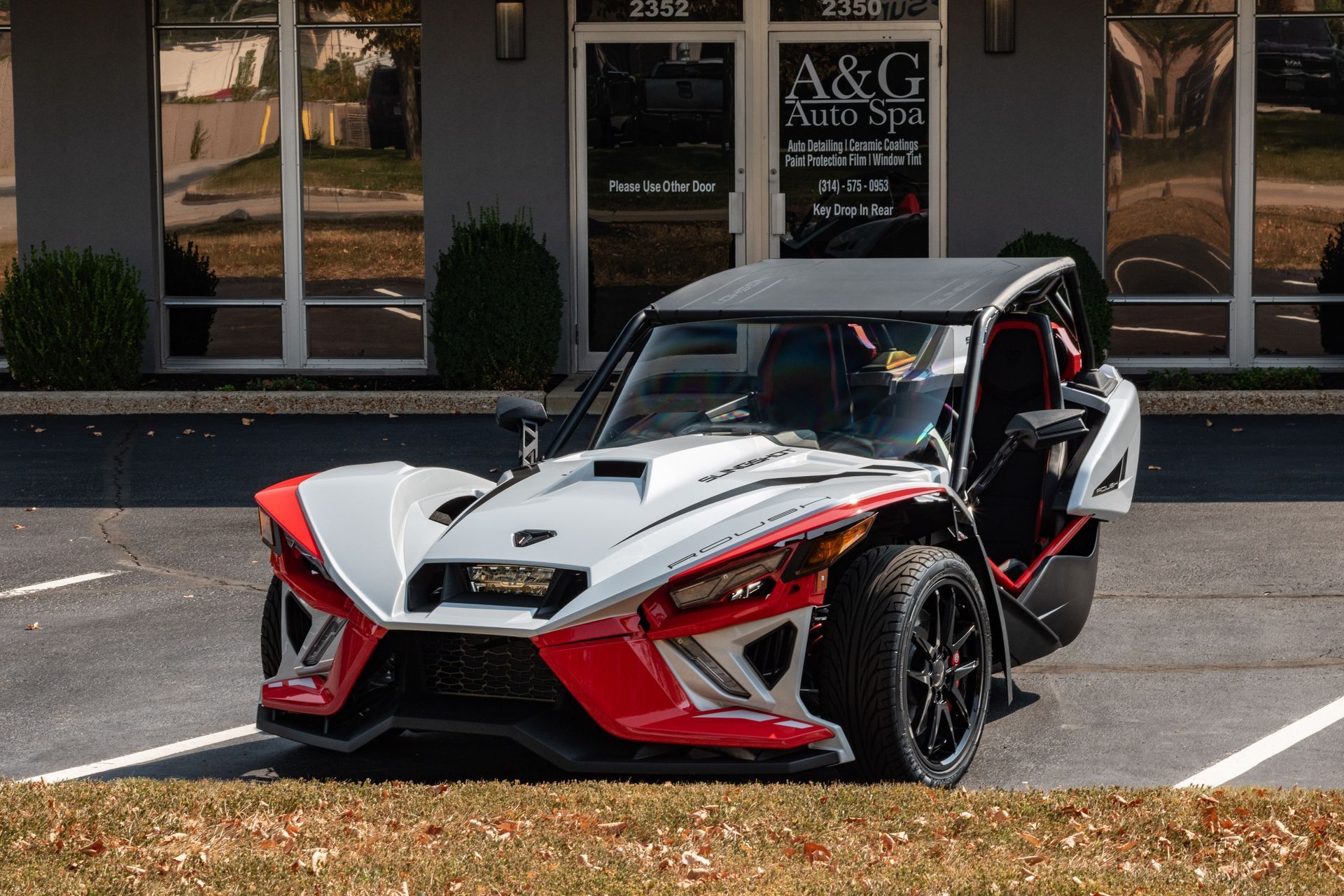 A red and white slingshot is parked in a parking lot in front of a building.