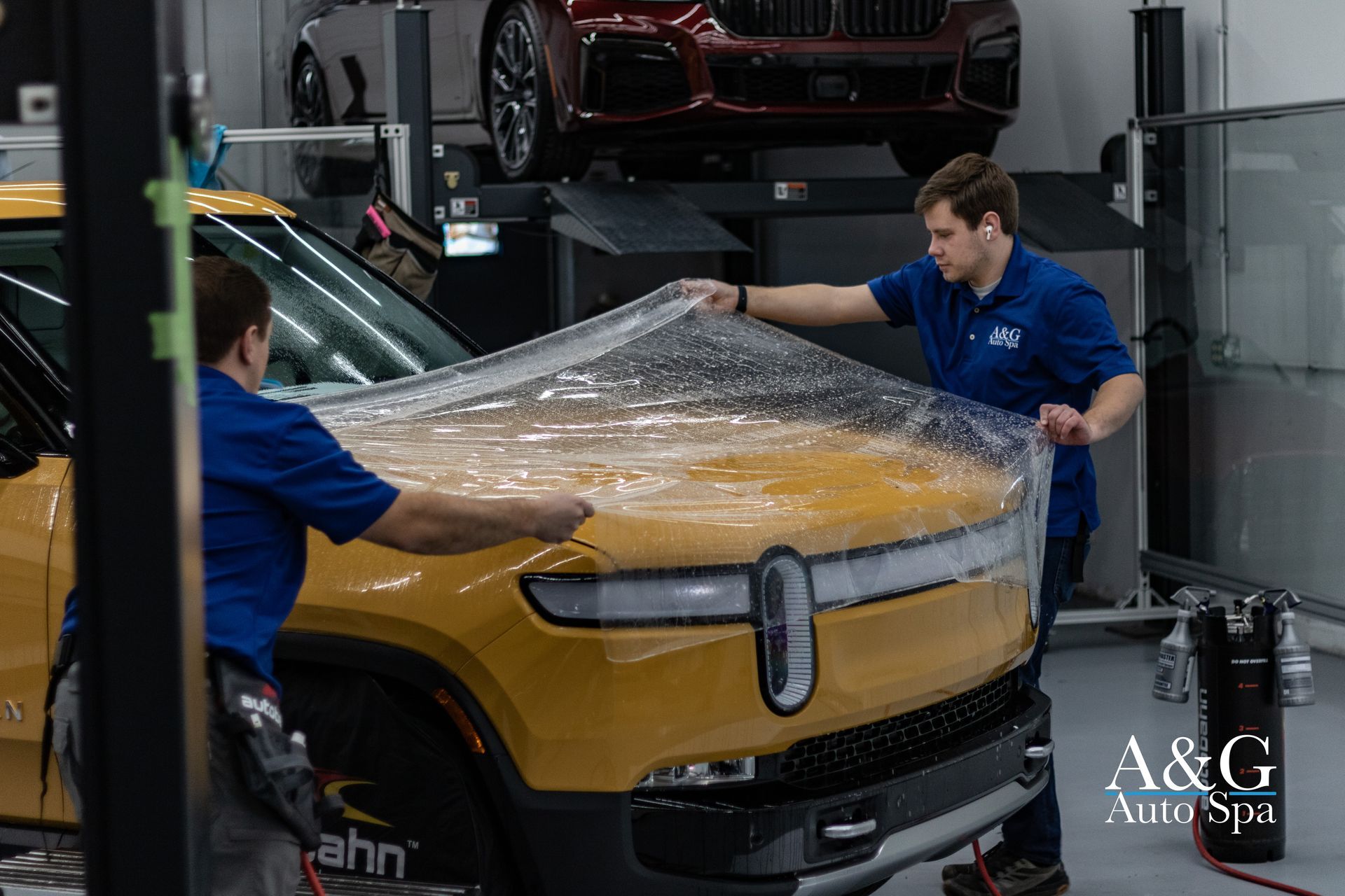two men are applying paint protection film to a yellow rivian R1T with plastic wrap in a garage.