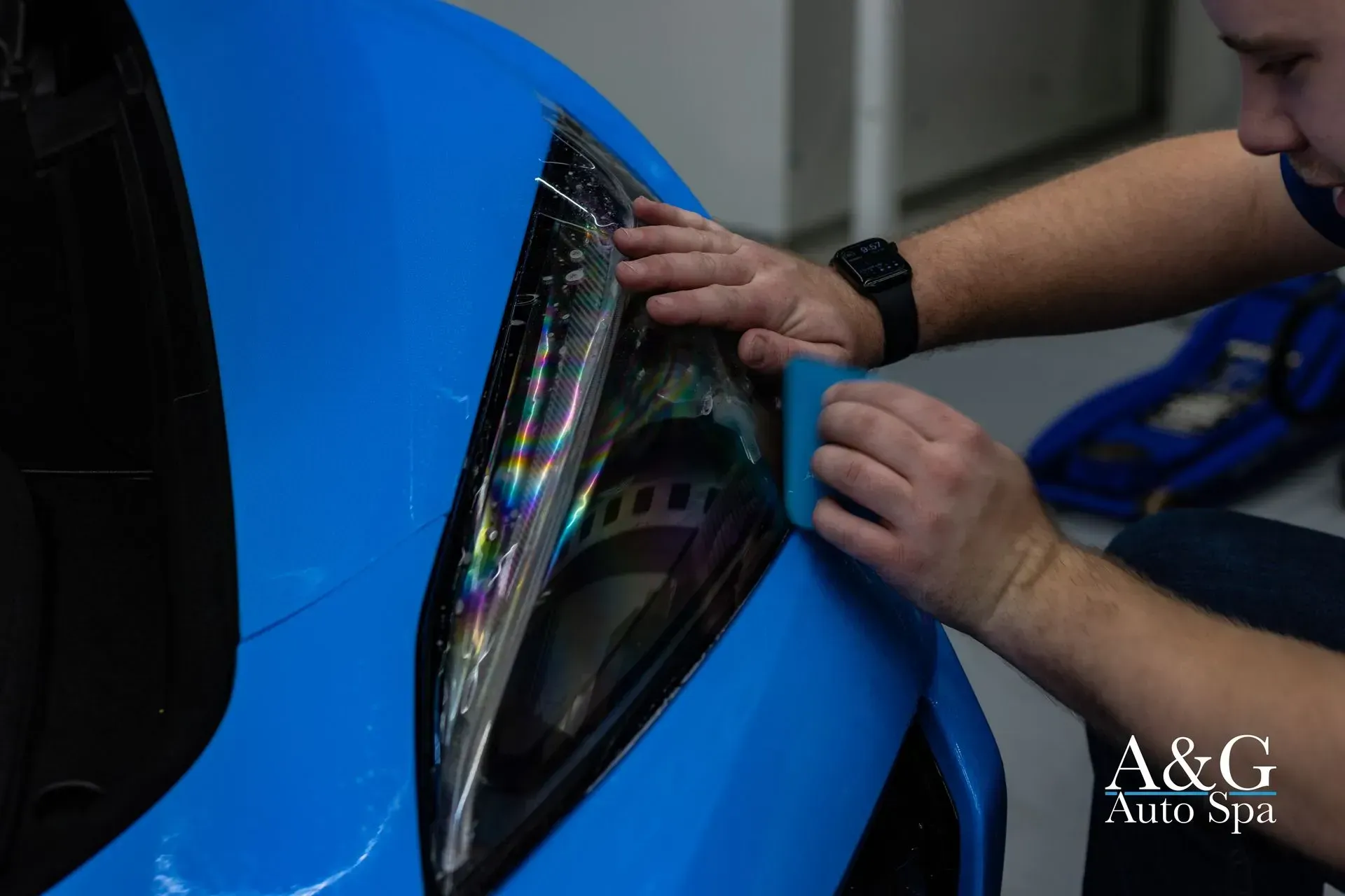 A man is applying a film to the headlight of a blue car.
