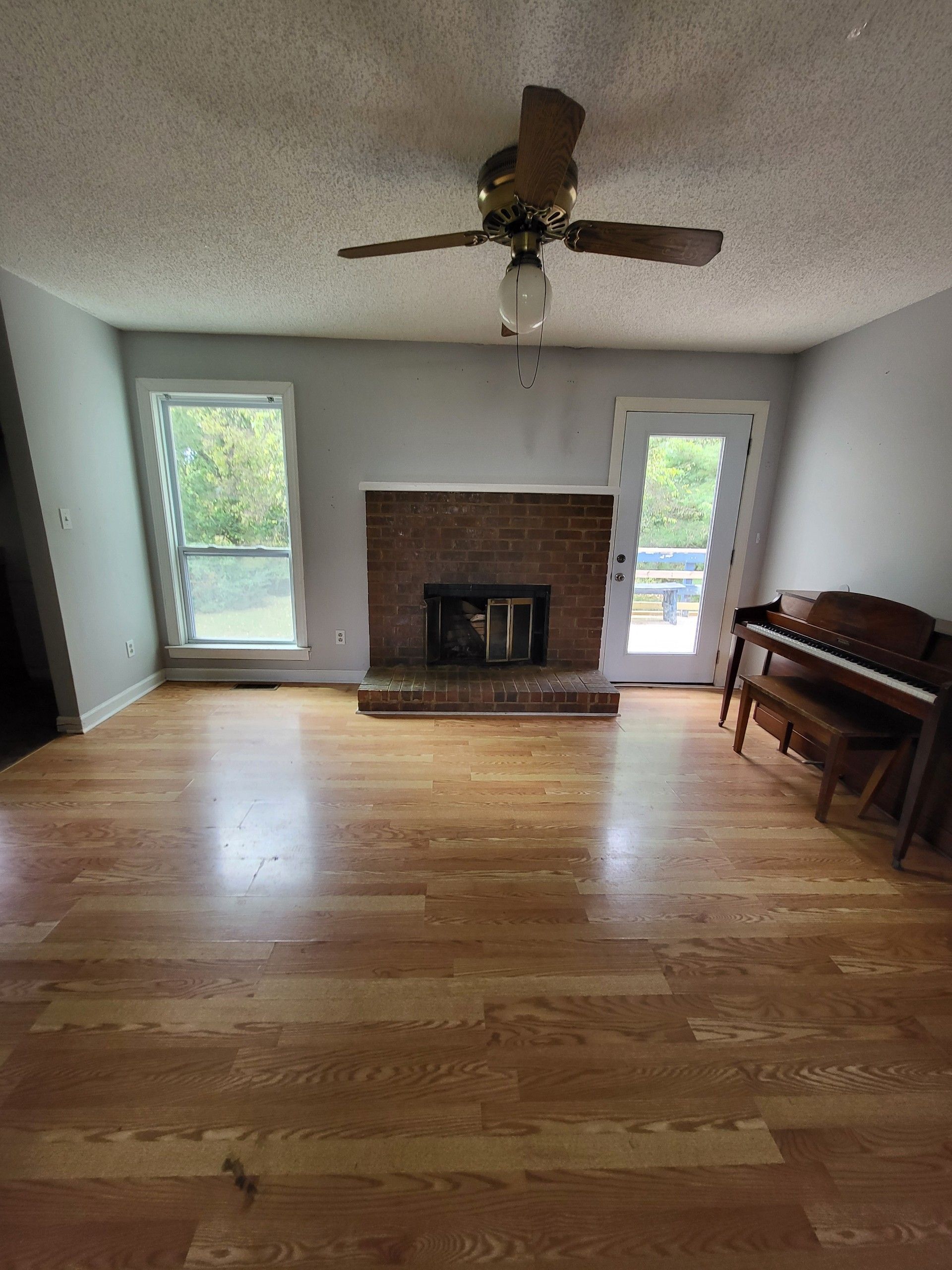 Living room with hardwood floors, a brick fireplace, and a piano. Gray walls, a ceiling fan, and windows are visible.