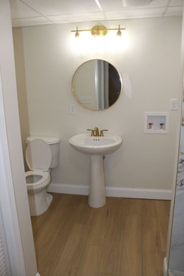 Bathroom with toilet, pedestal sink, round mirror, and gold fixtures; neutral walls and wood-look floor.