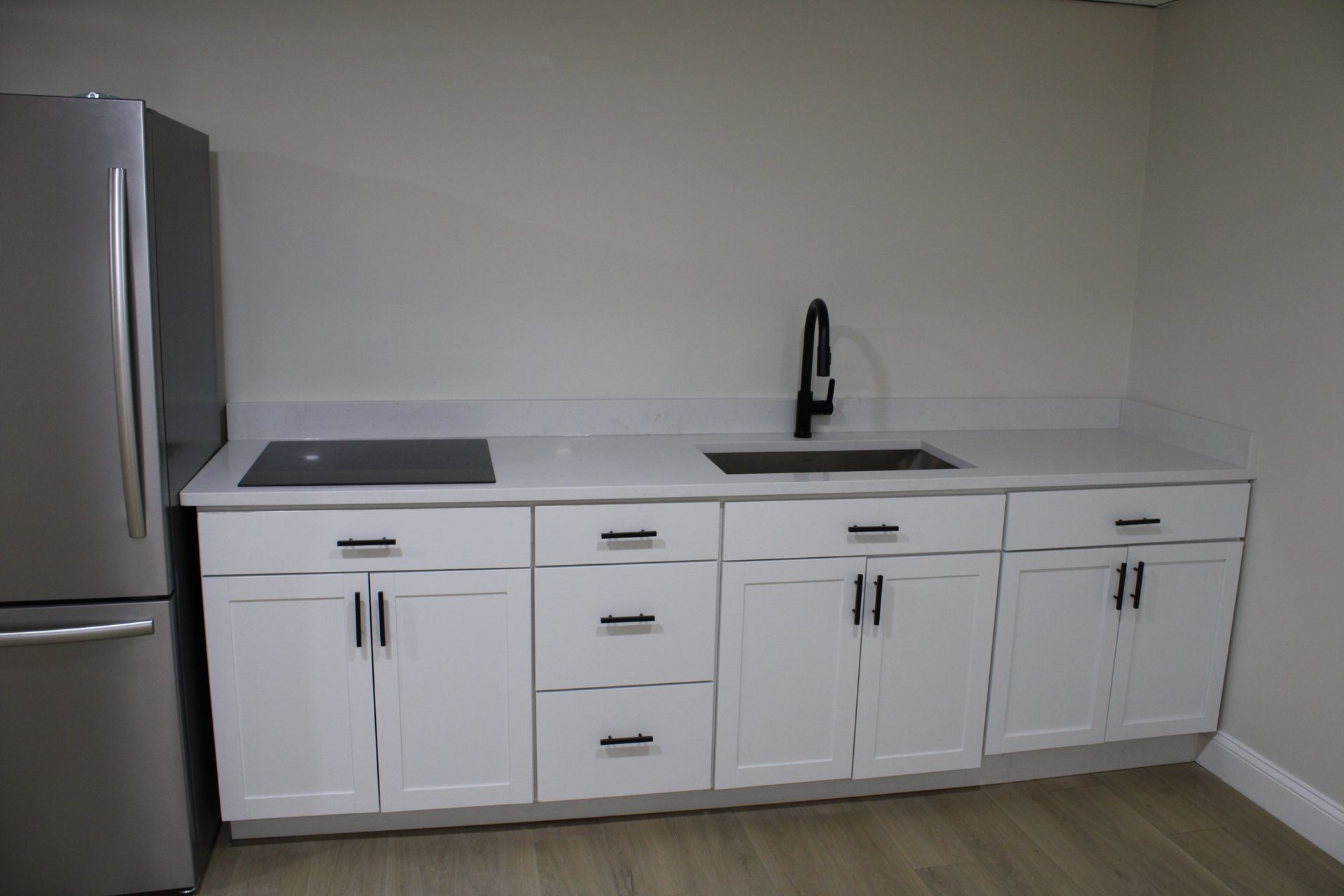 White kitchen cabinets with black hardware, sink, and cooktop. Stainless steel refrigerator.