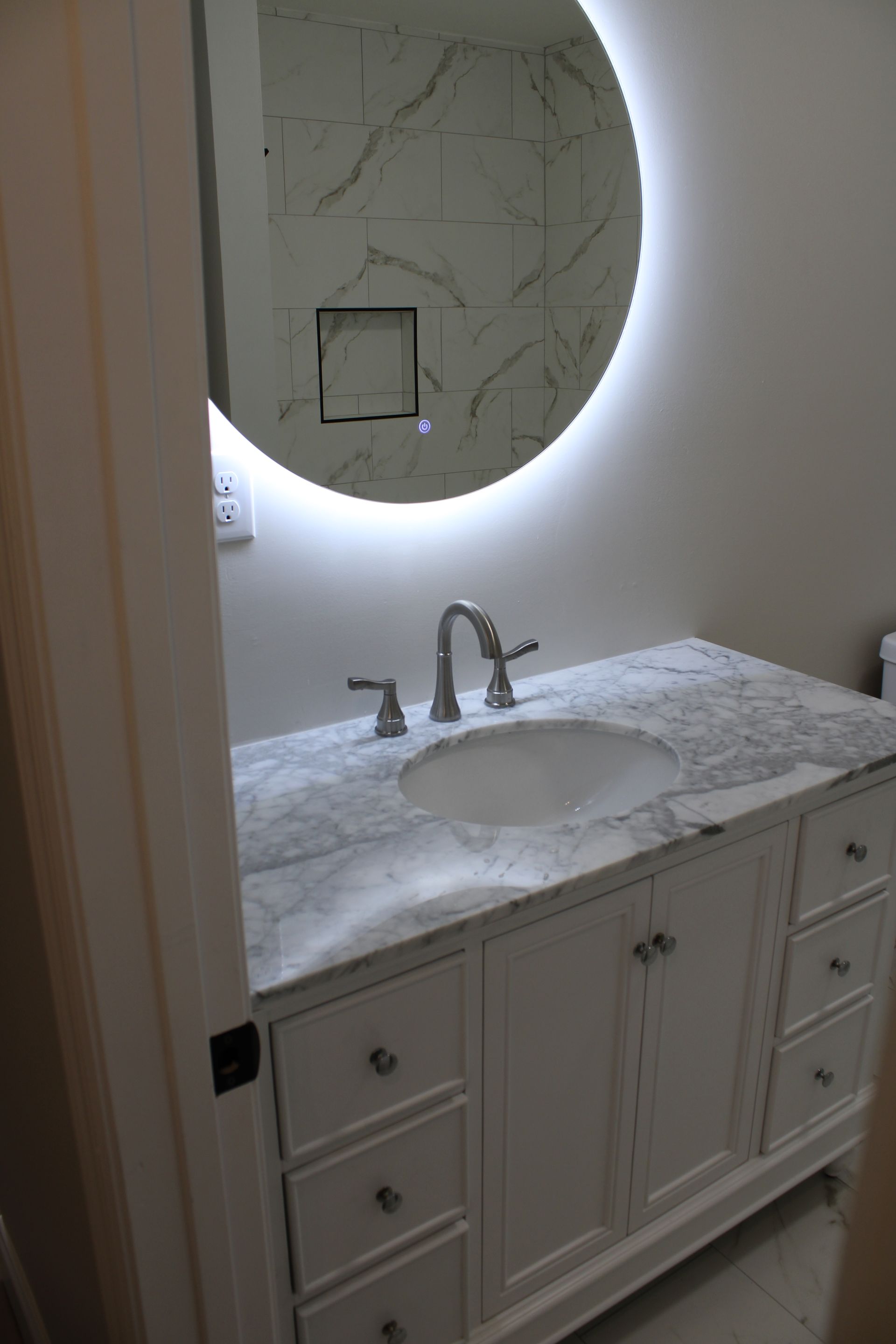 White bathroom vanity with marble countertop, round backlit mirror, and silver faucet.