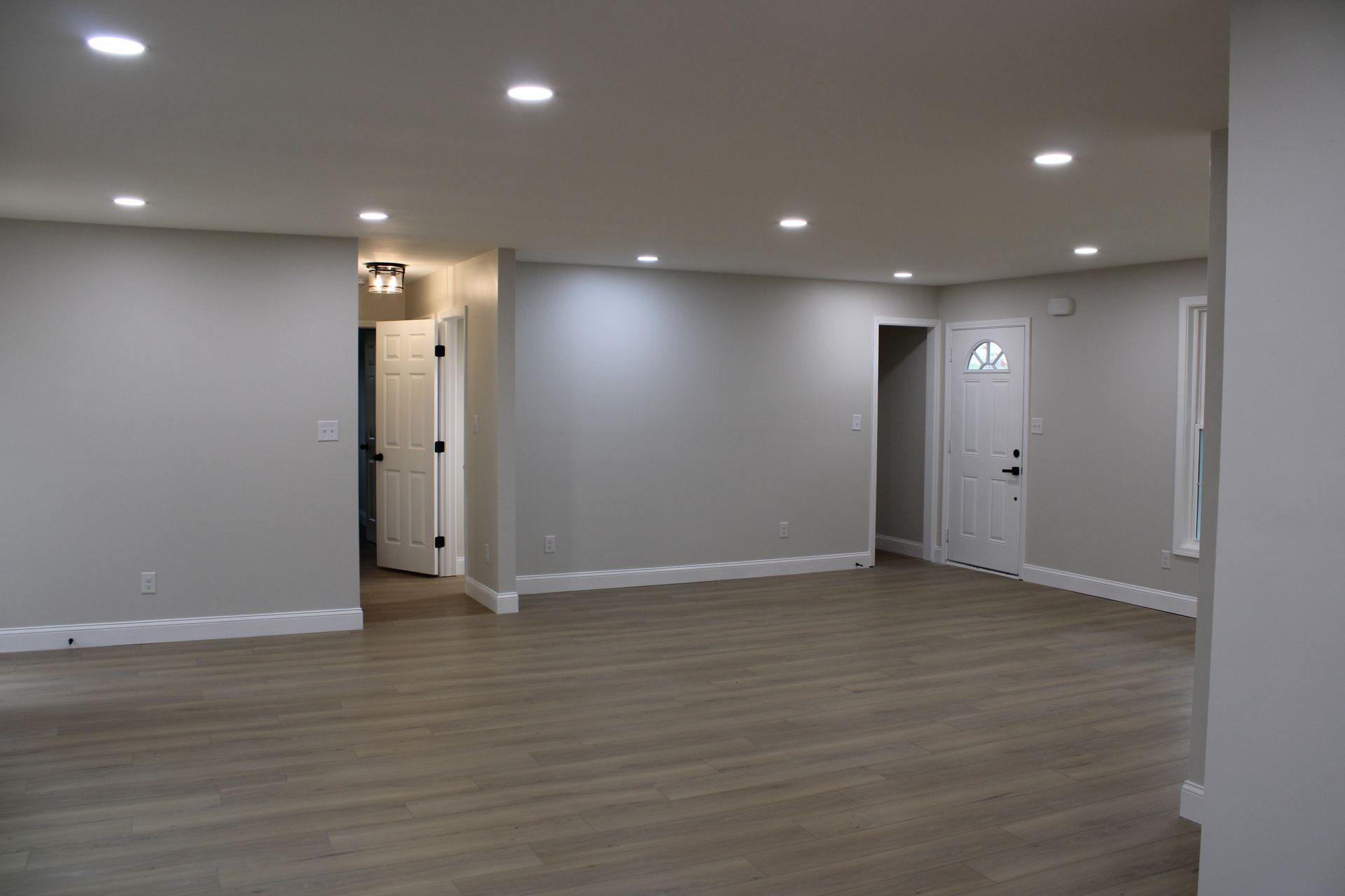Empty, freshly painted living room with recessed lighting and wood-look flooring. Doors lead to other rooms.