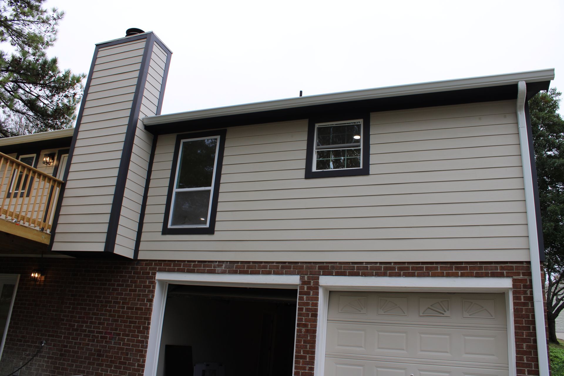 Tan house with brick base and two-car garage. Windows, chimney, and a wooden balcony are visible.