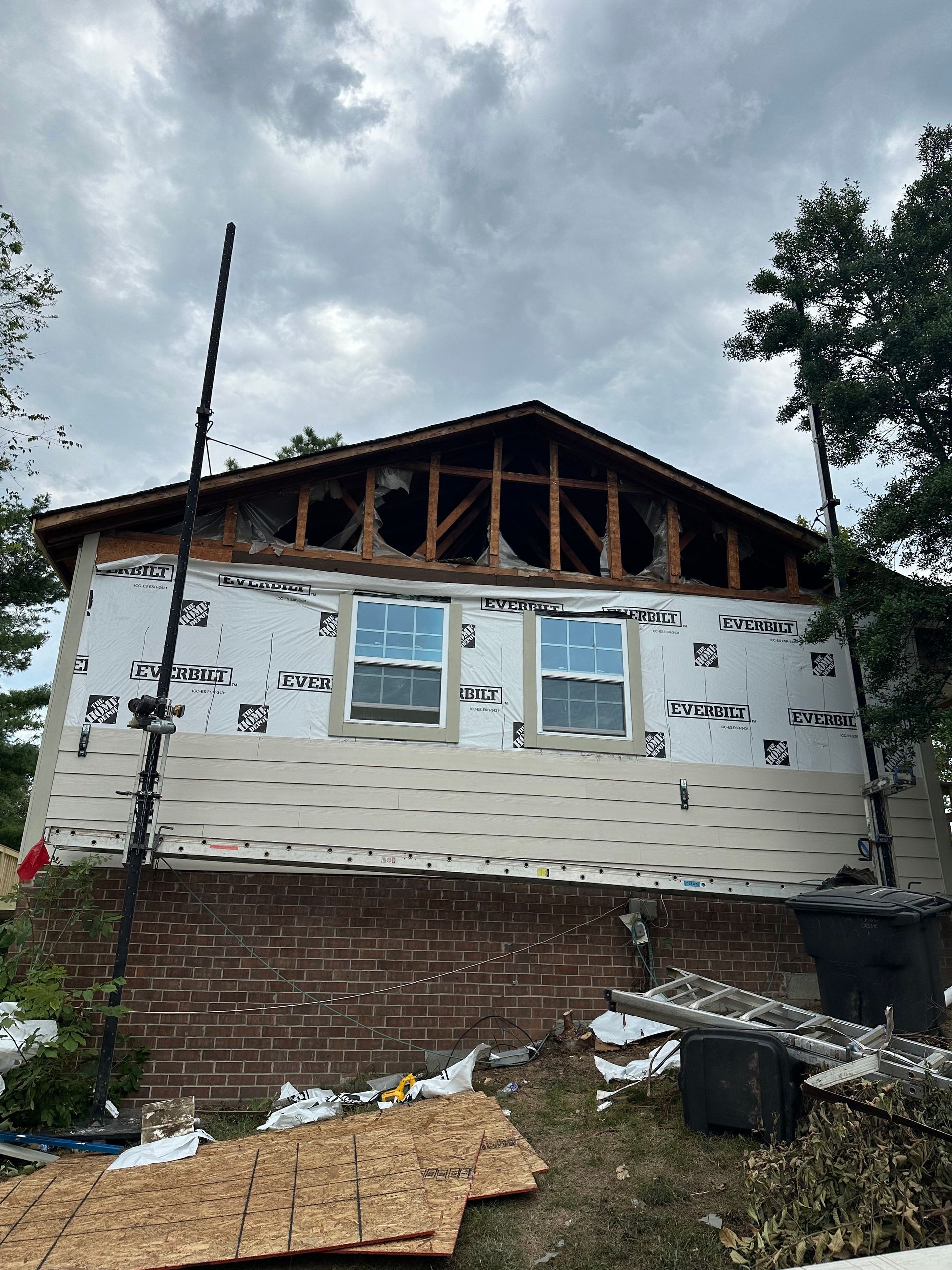 House under construction: damaged roof, exposed wood framing, two windows, brick foundation, overcast sky.