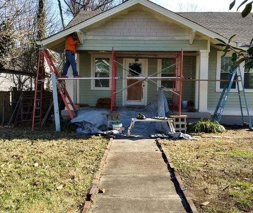 Man on a scaffold painting the front porch of a house. Scaffolding, ladders, and tarps are visible.