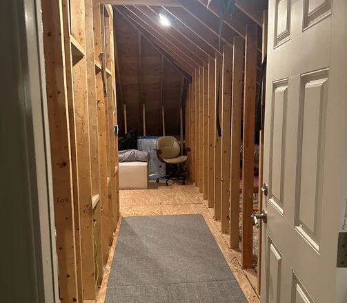 Hallway in an unfinished attic space with exposed wood framing, and a rug. Doorway in the foreground.