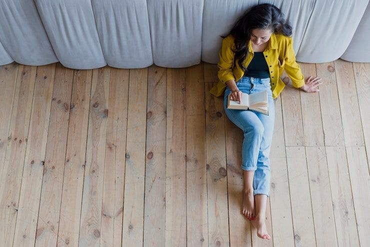 Woman in yellow shirt and jeans reads a book on a wooden floor, leaning against a bed.