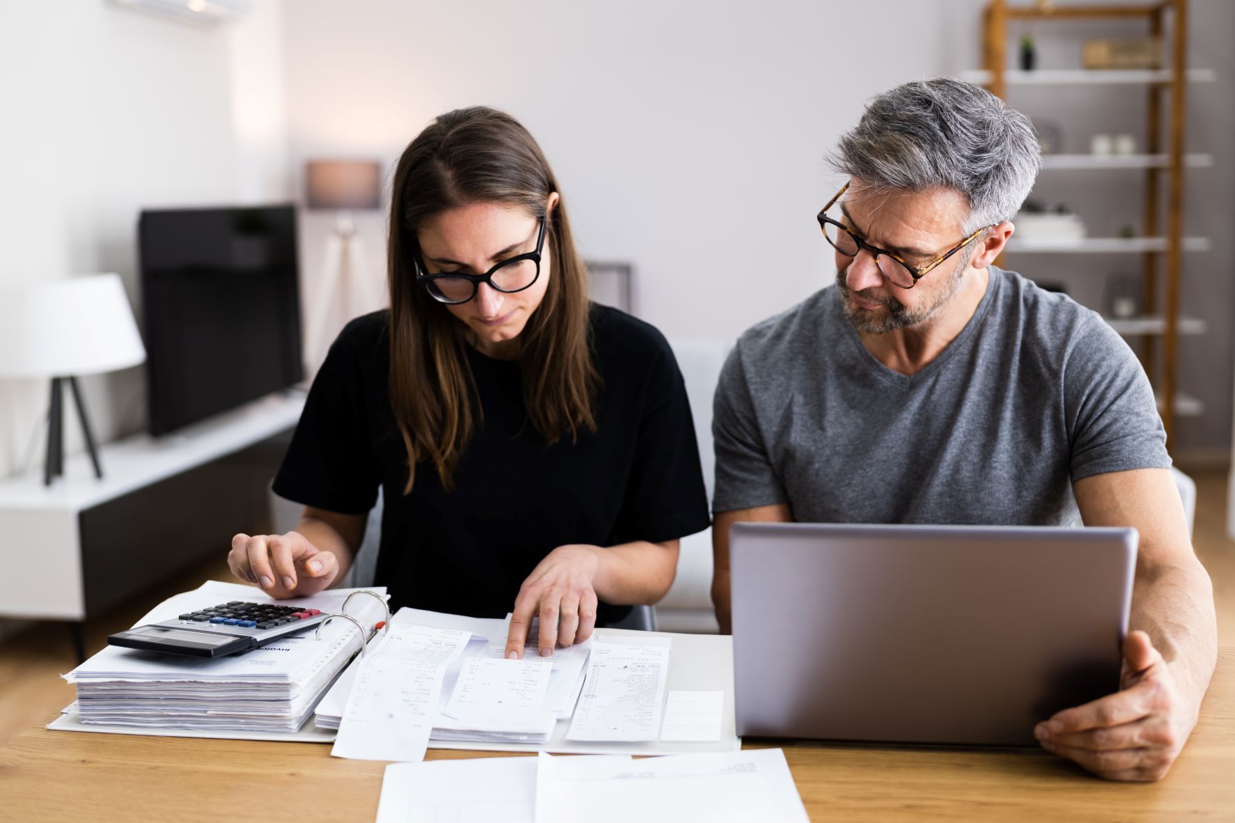 Two adults sit at a table reviewing receipts, paperwork, and computer together.