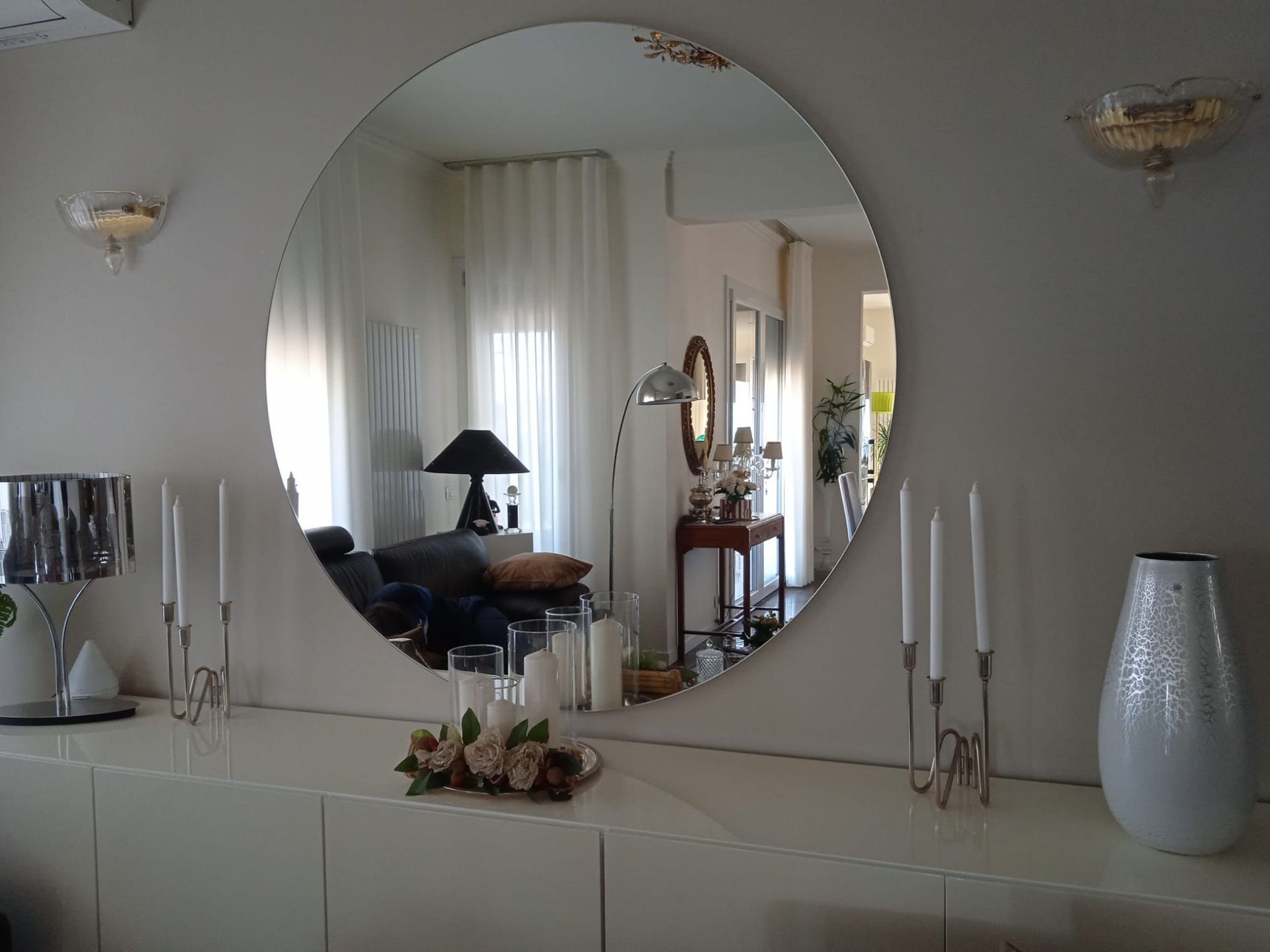 In un soggiorno, sopra una credenza bianca con portacandele, lampade e un vaso, è appeso uno specchio rotondo a parete.