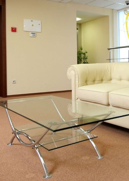 Un tavolino da caffè con piano in vetro e struttura in metallo, posizionato di fronte a un divano in pelle bianca trapuntata, nella hall di un moderno ufficio.