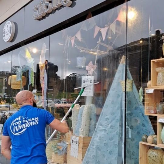 A man wearing a blue shirt that says clean windows is cleaning a store window