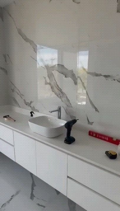 Baño moderno blanco con pared con diseño de mármol, tocador blanco, lavabo sobre encimera y un taladro negro.