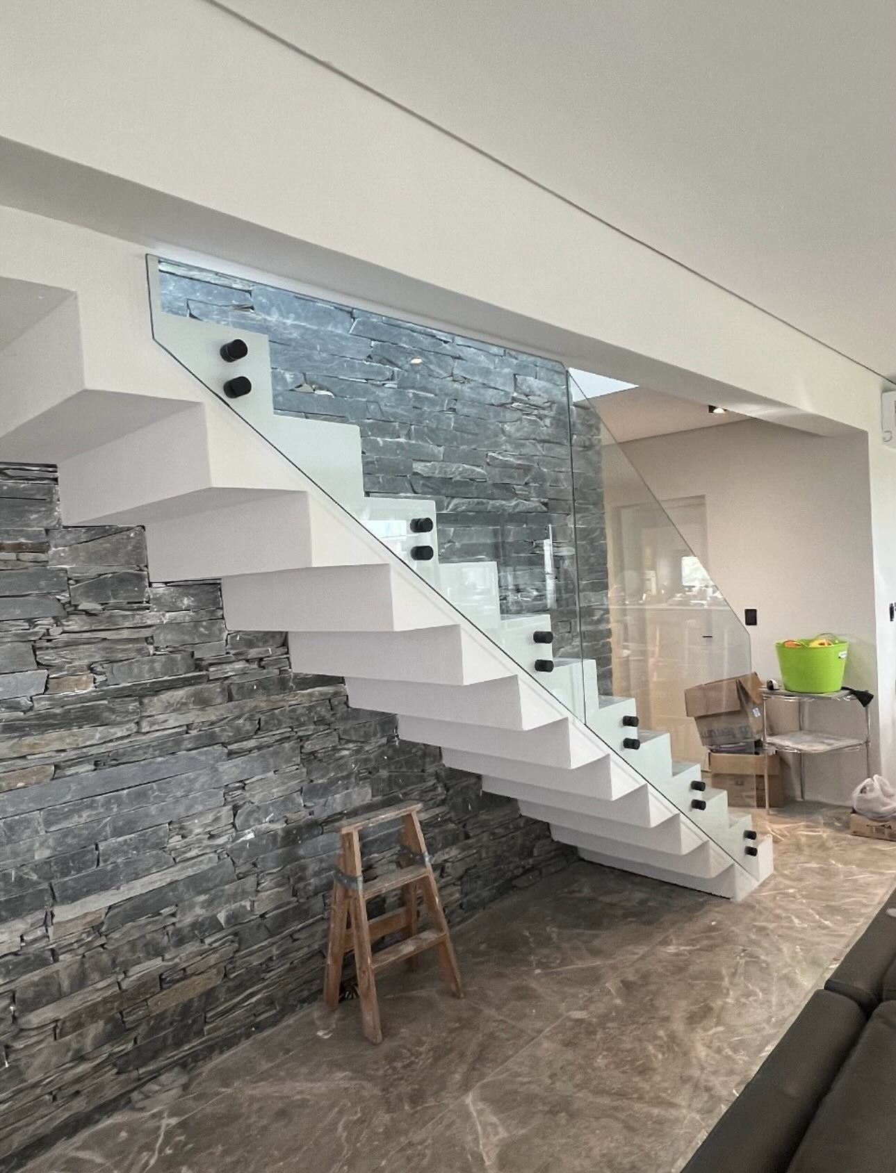 Escalera blanca moderna con barandilla de cristal. Muro de piedra a la izquierda. Pequeña escalera de madera.