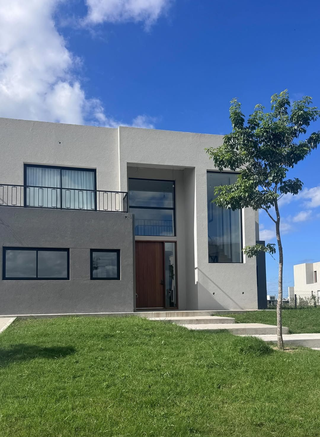 Casa gris moderna con grandes ventanales, puerta de madera y un pequeño árbol en un césped verde bajo un cielo azul.
