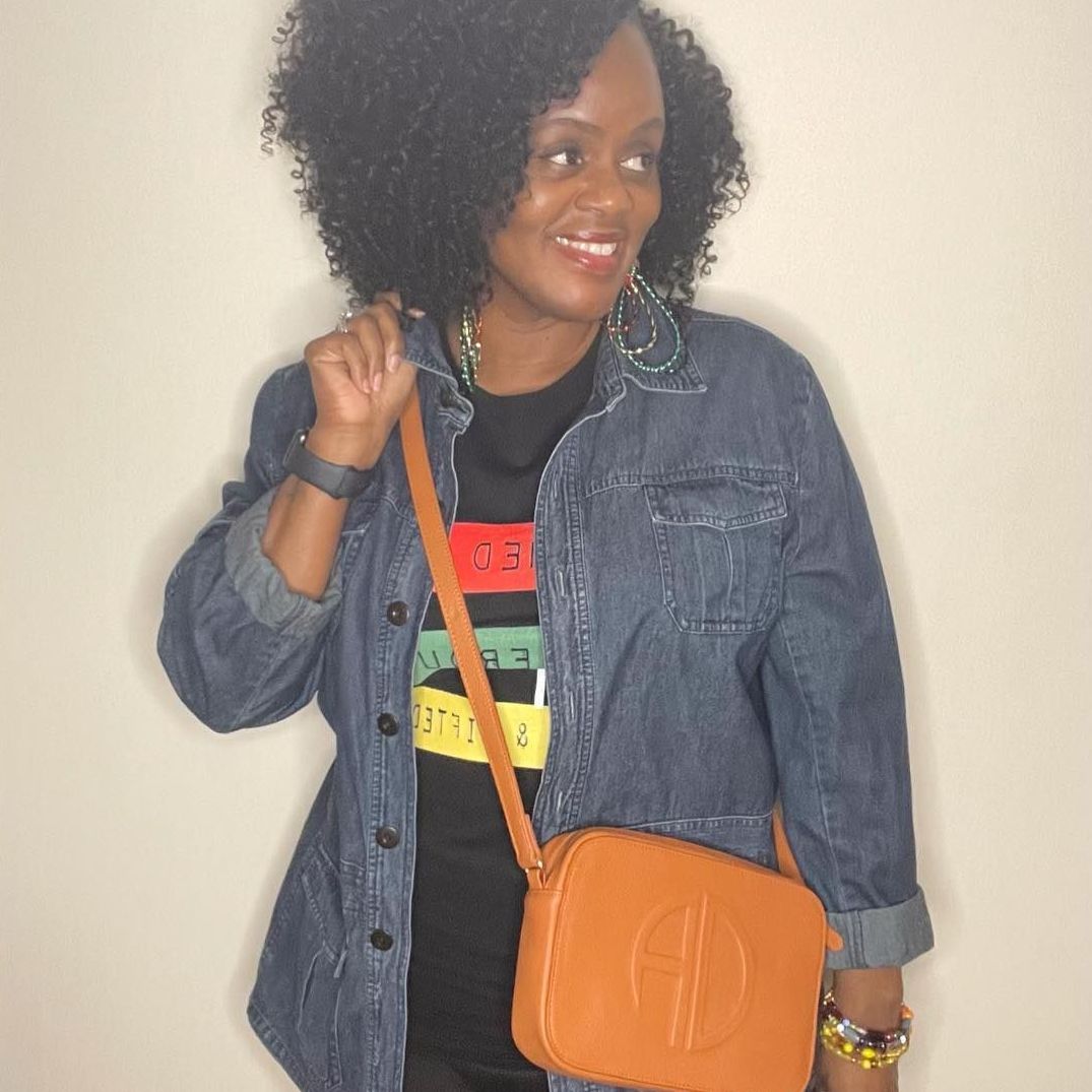 Woman with curly hair, in denim jacket, black shirt with colorful stripes, carrying orange purse, smiling.