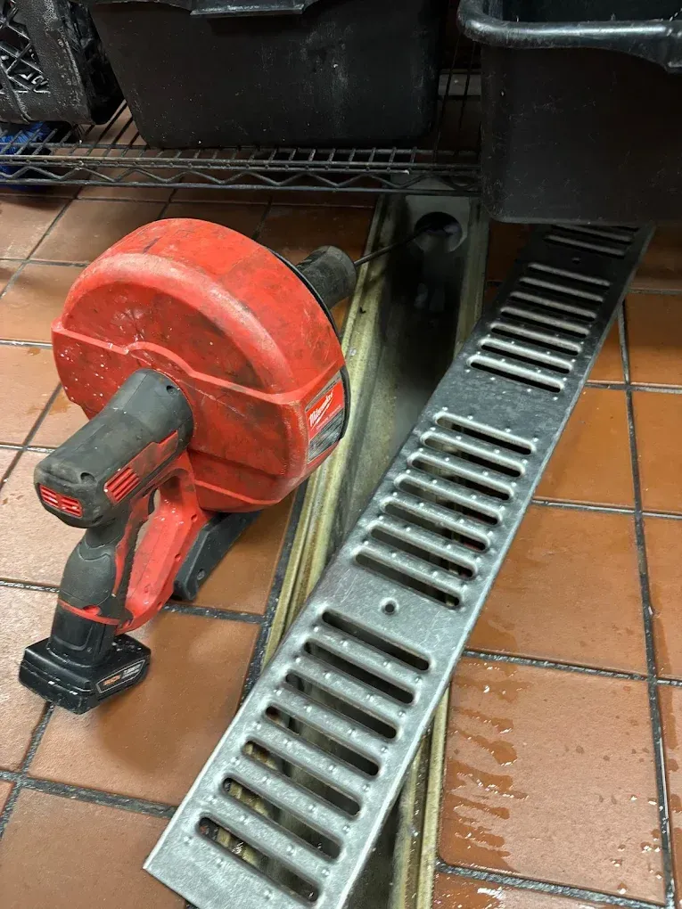 Red and black drain snake tool next to a metal drain in a commercial kitchen setting.