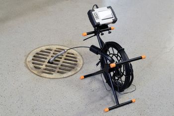 Drain inspection camera set up over a storm drain. A black reel and stand support a camera with a monitor.