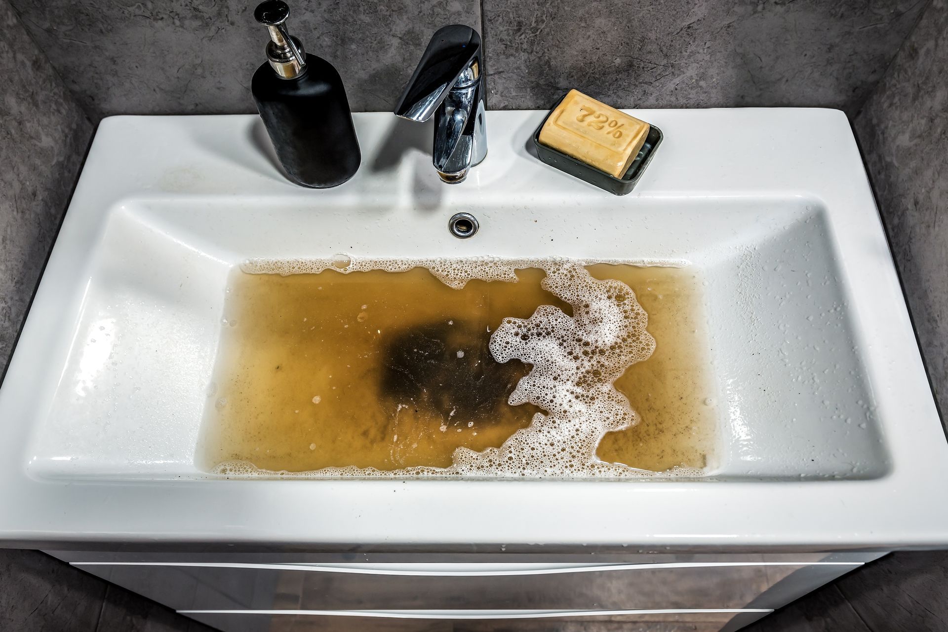 A white bathroom sink filled with murky brown water, soap, and a soap dispenser.
