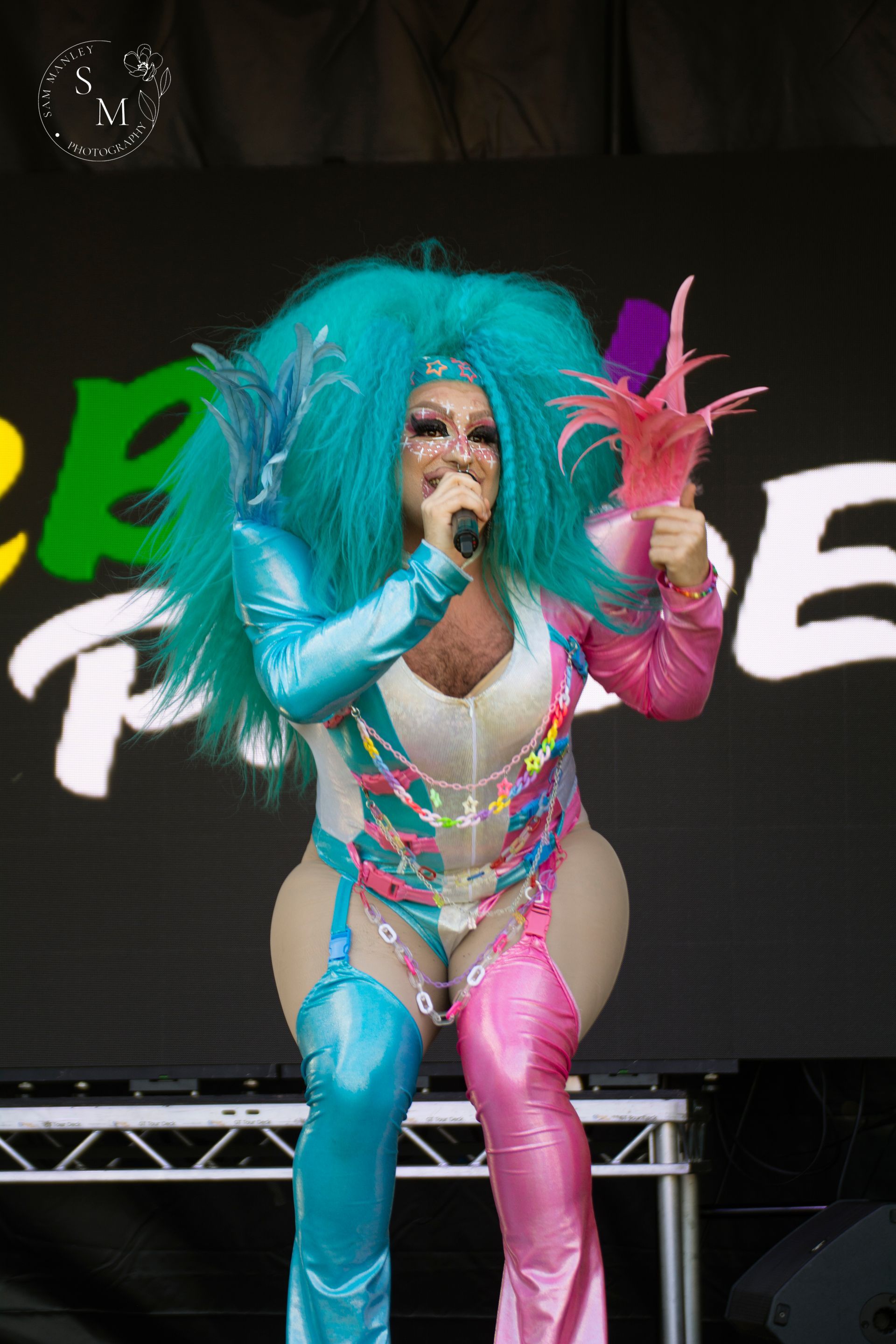 Drag queen Felica Blueball performing on stage with bright turquoise hair, colorful makeup, and a blue and pink costume.