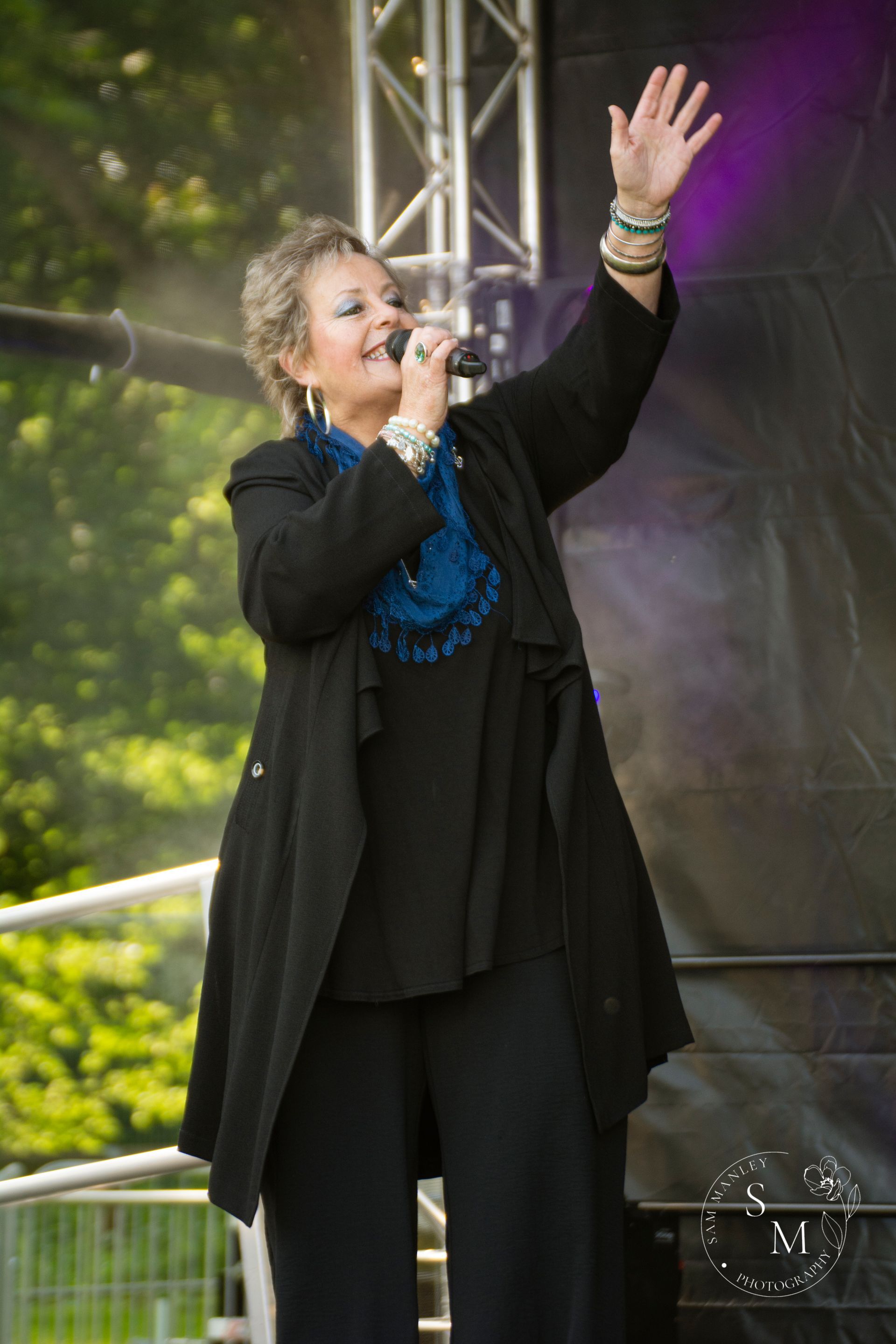 Woman singing on stage with a microphone, wearing black clothing and a blue scarf, raising one hand.