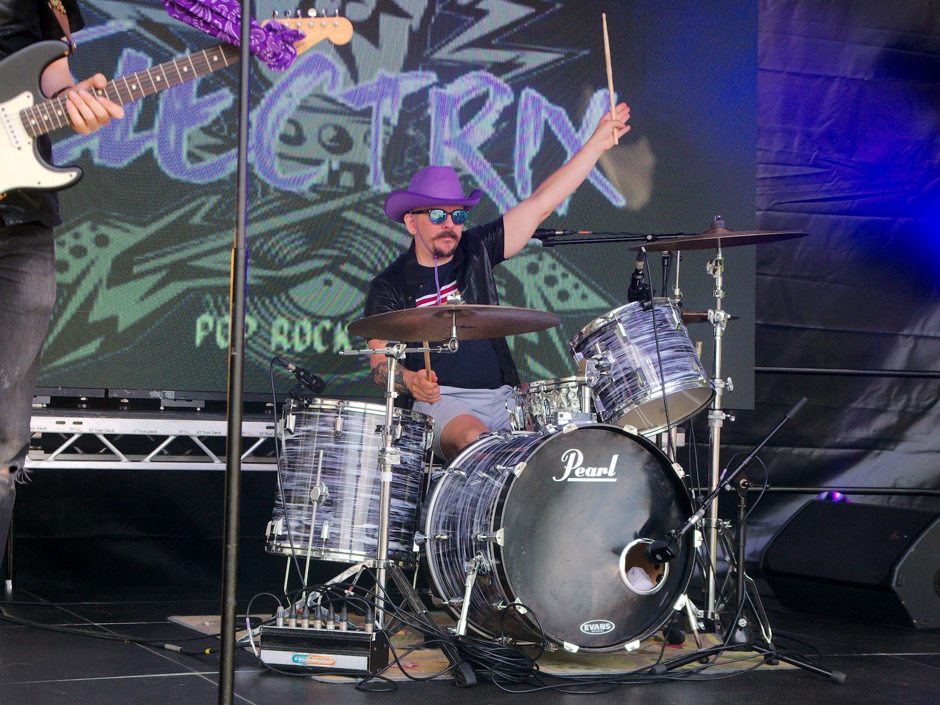 A Drummer playing on stage, wearing a purple hat and sunglasses, holding one drumstick in the air.