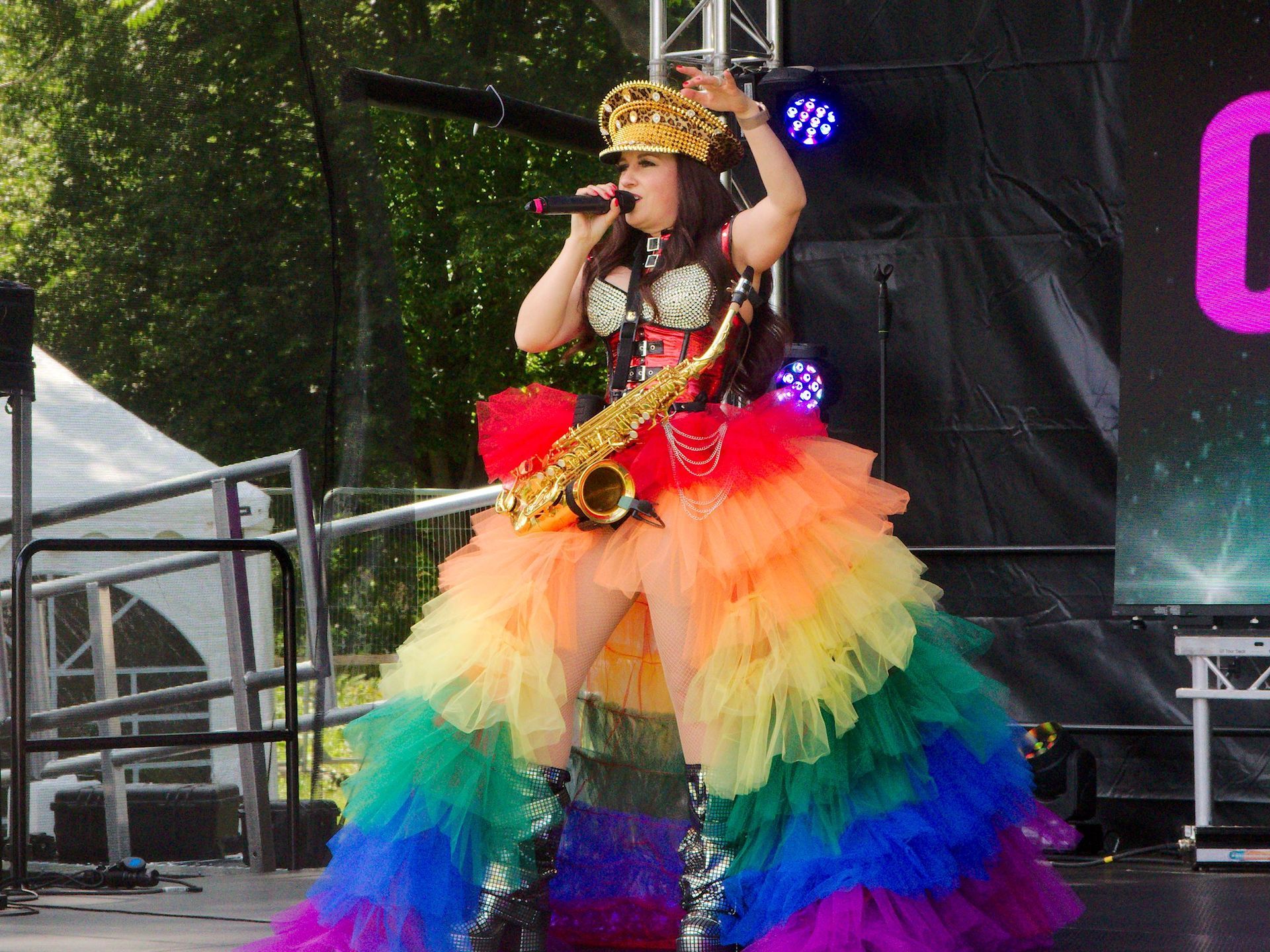 A Performer singing on stage with a microphone, wearing a rainbow dress, gold crown, and holding a saxophone.