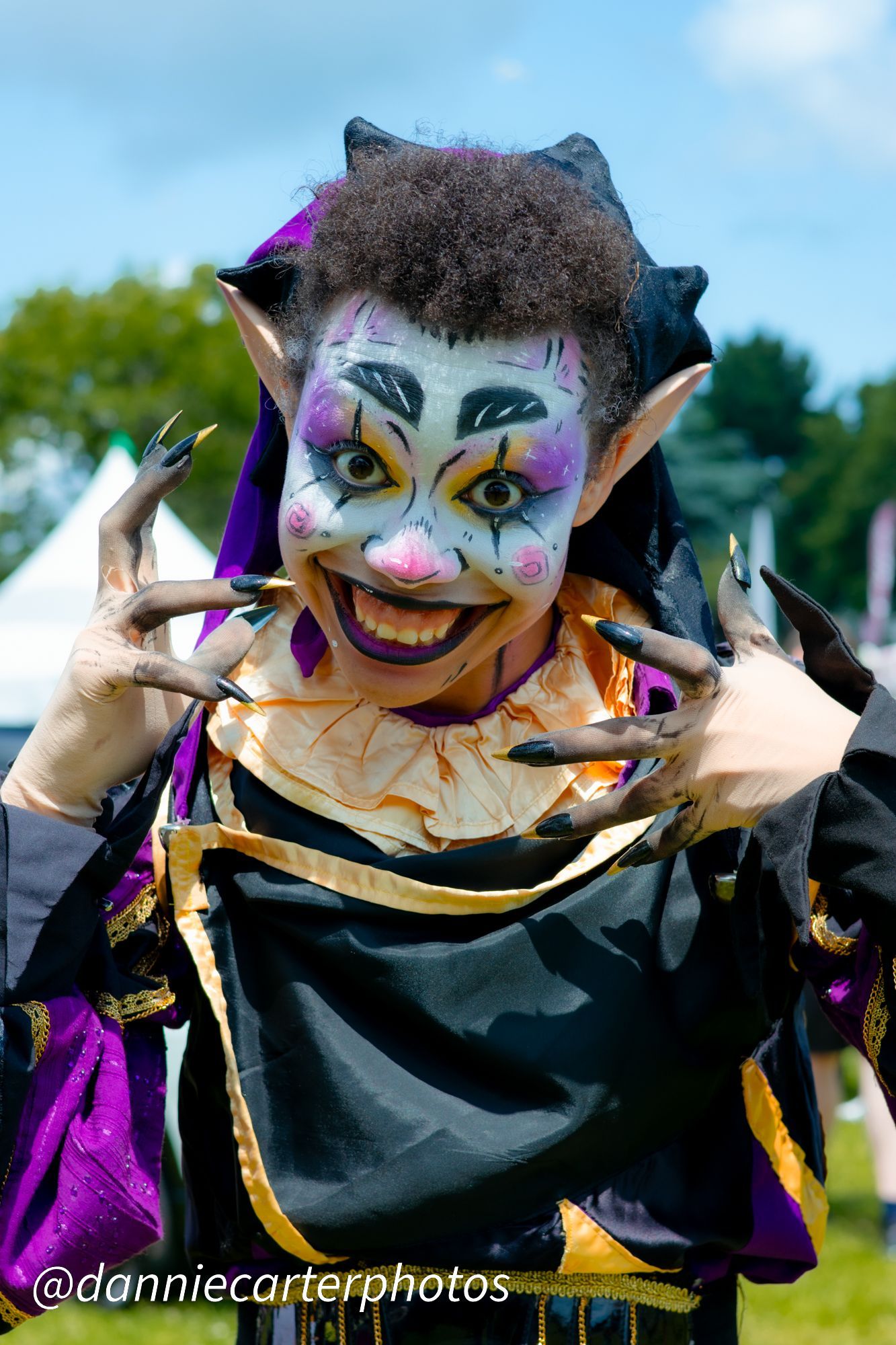 Drag jester Razzle B Dazzled in colorful costume and face paint, posing with clawed hands and a wide smile.