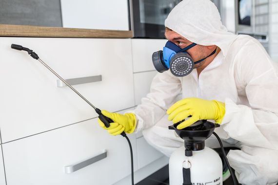 Pest control worker spraying white cabinets in a kitchen, wearing protective gear.