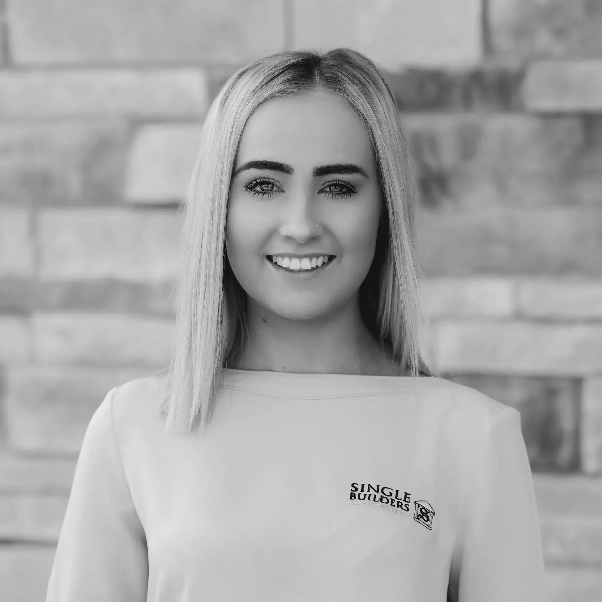 A black and white photo of a woman smiling in front of a brick wall.