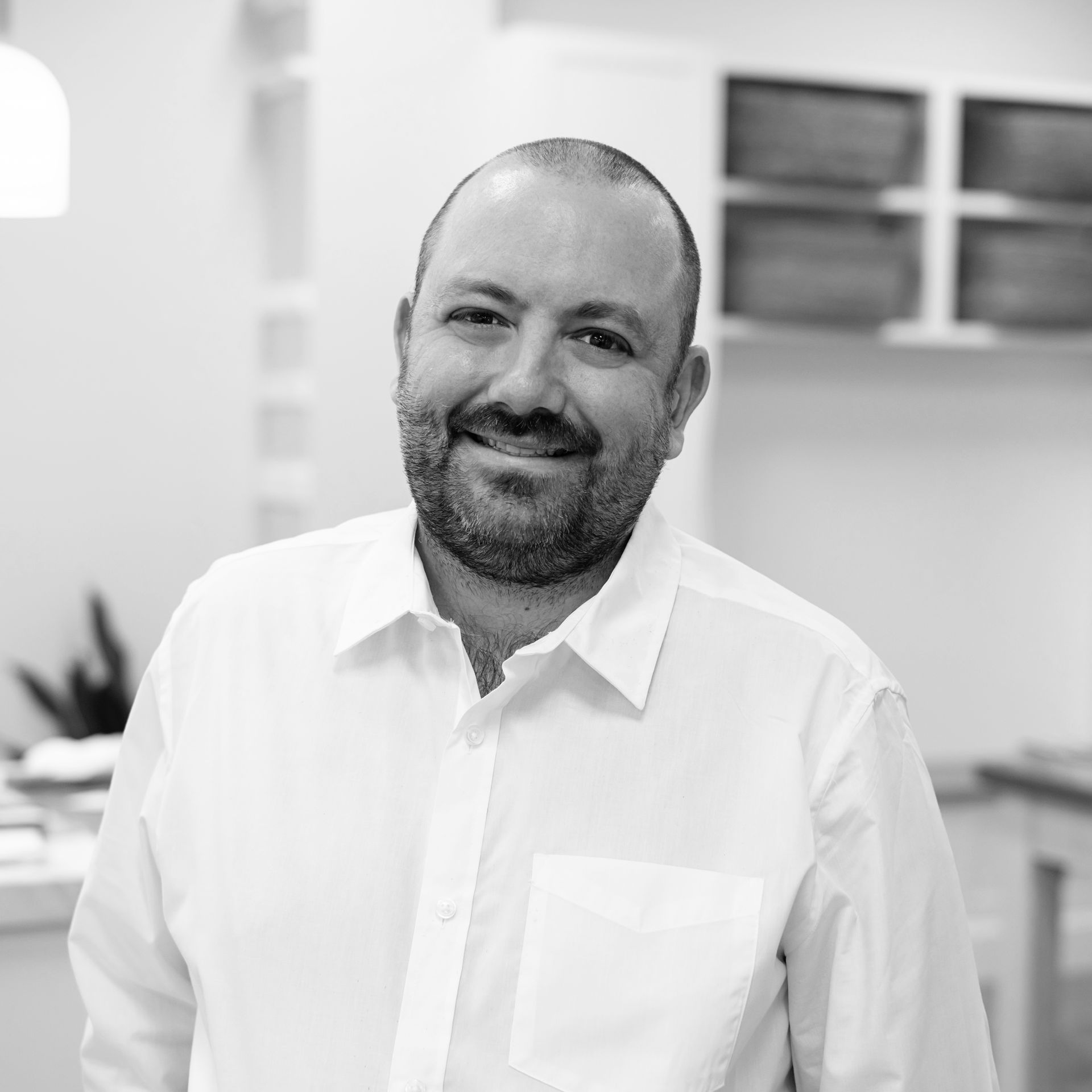 A man with a beard is wearing a white shirt and smiling in a black and white photo.